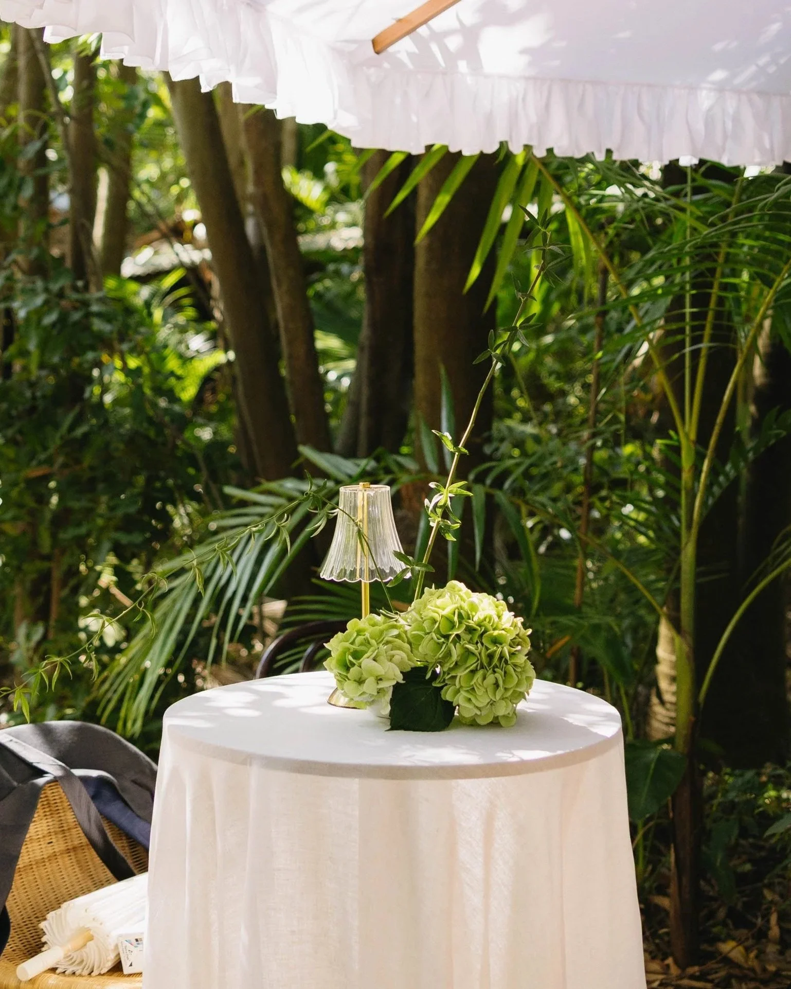 A small table with a white tablecloth is outdoors surrounded by green foliage. On the table is a floral arrangement of light green hydrangeas with a small white lampshade. A white ruffled parasol provides partial shade above.