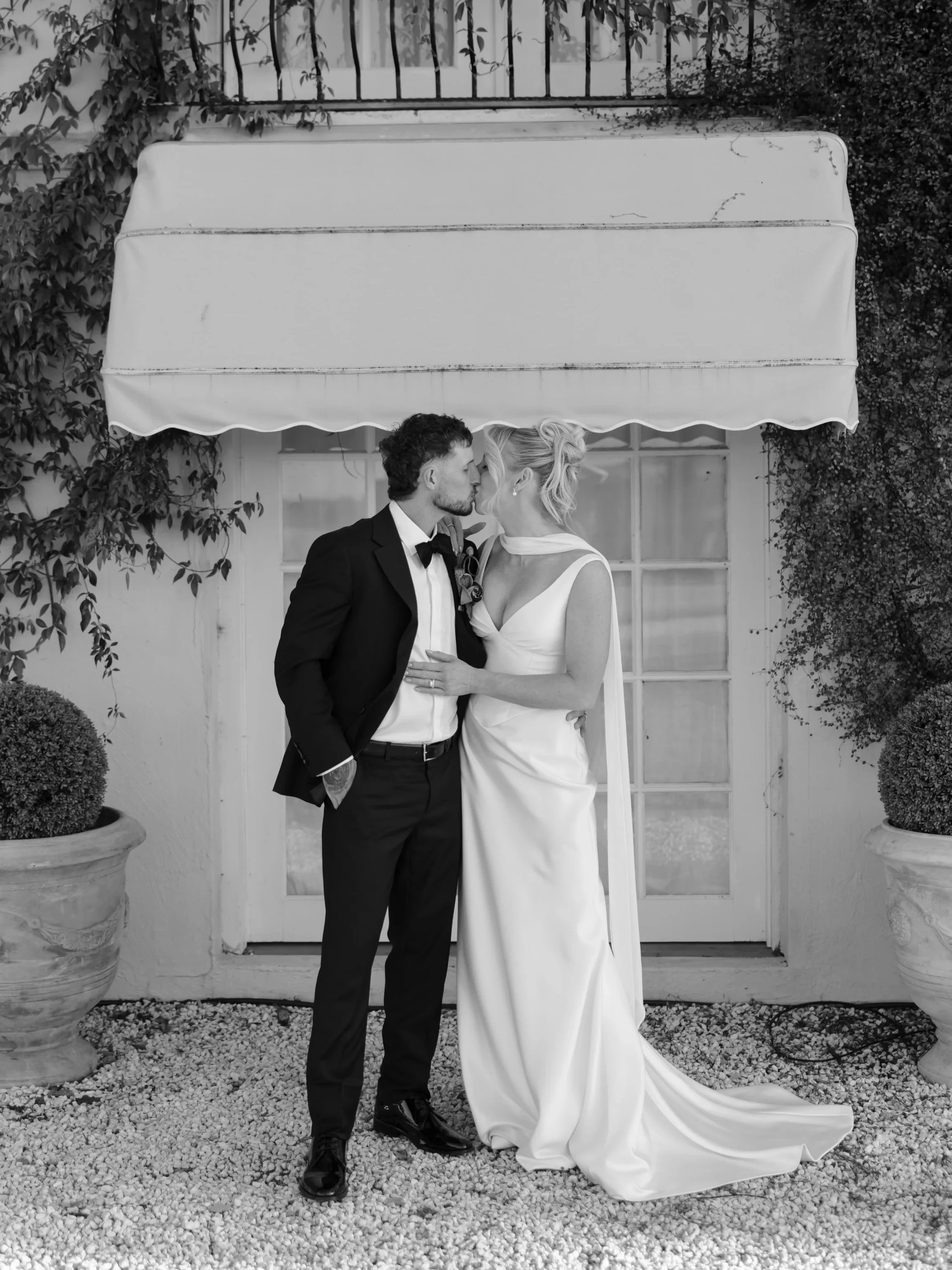 A black and white photo of a bride and groom kissing in front of a white door with glass panes, surrounded by potted plants and greenery.