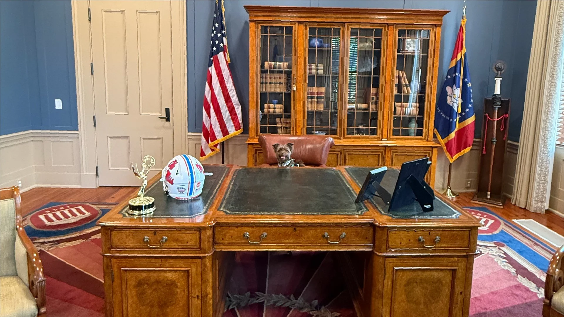 Office with wooden desk, flagpoles with American and state flags, a glass-front bookcase filled with books, and a small dog sitting behind the desk.