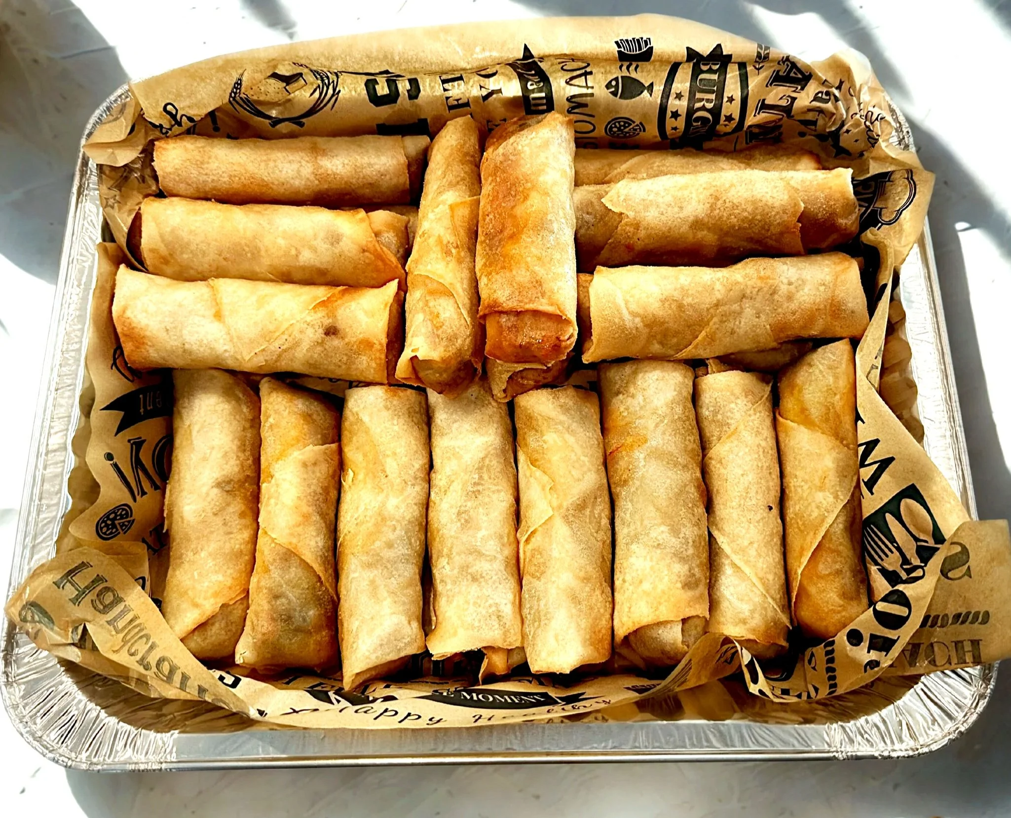 A tray of neatly arranged fried spring rolls in a foil container lined with printed parchment paper.