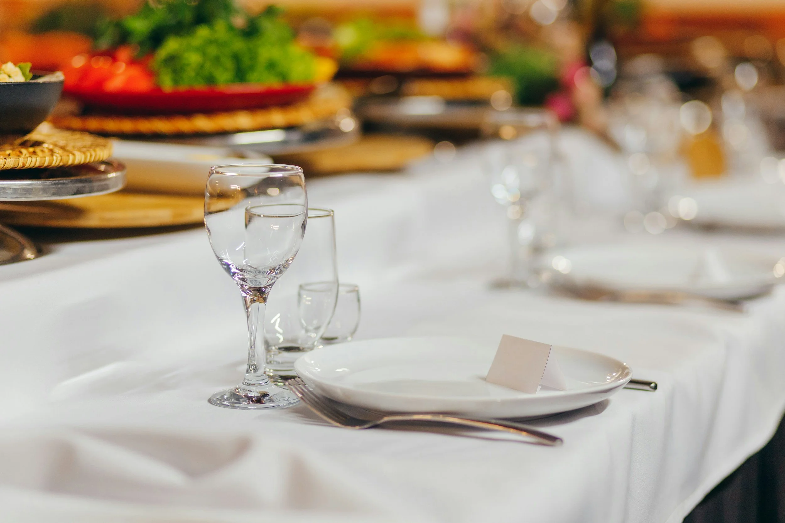Elegant banquet table setting with empty glassware, white plates, and a fork, with a blurred background of food and decorations.