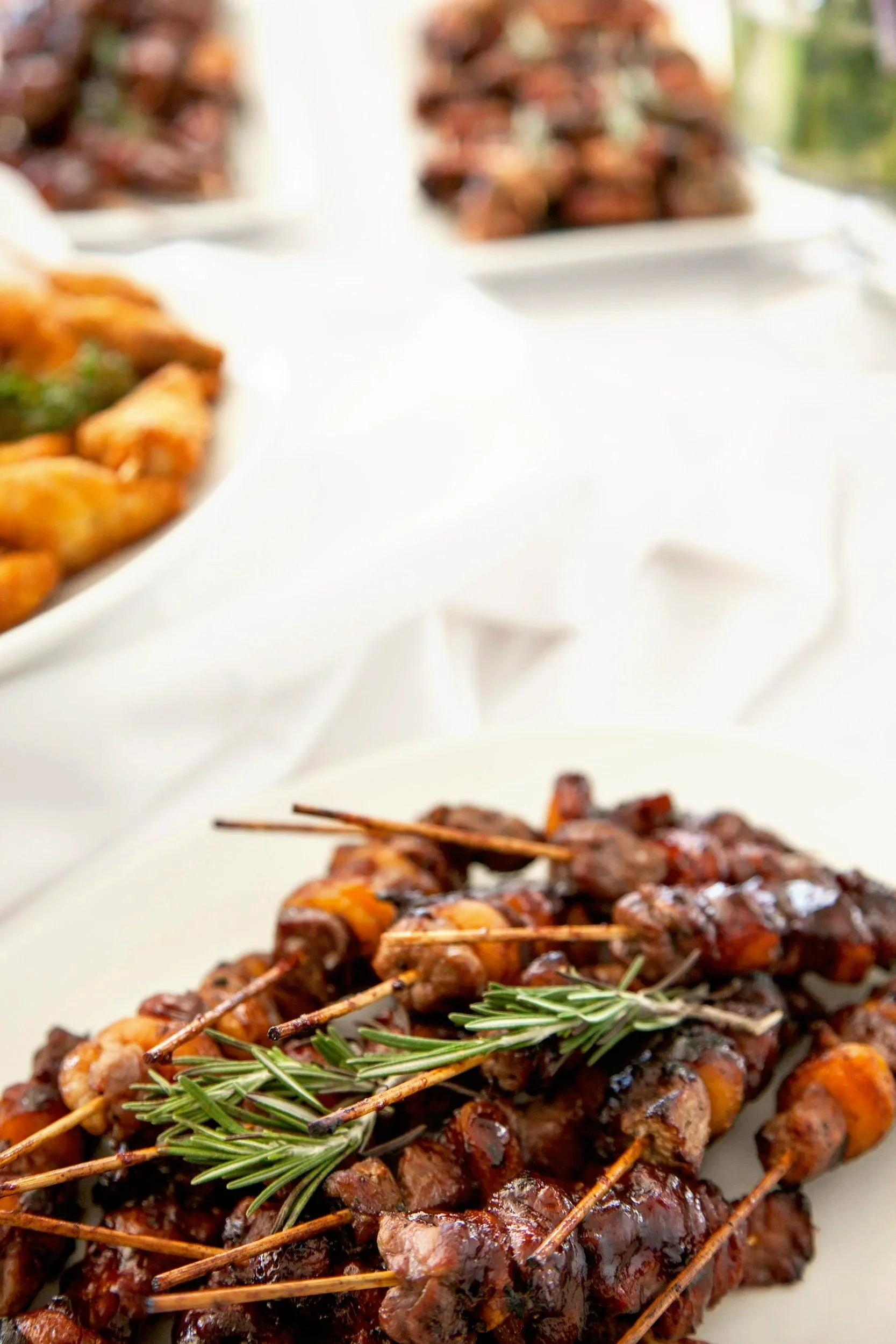 Close-up of grilled meat skewers garnished with a sprig of rosemary, on a white plate at a buffet table.