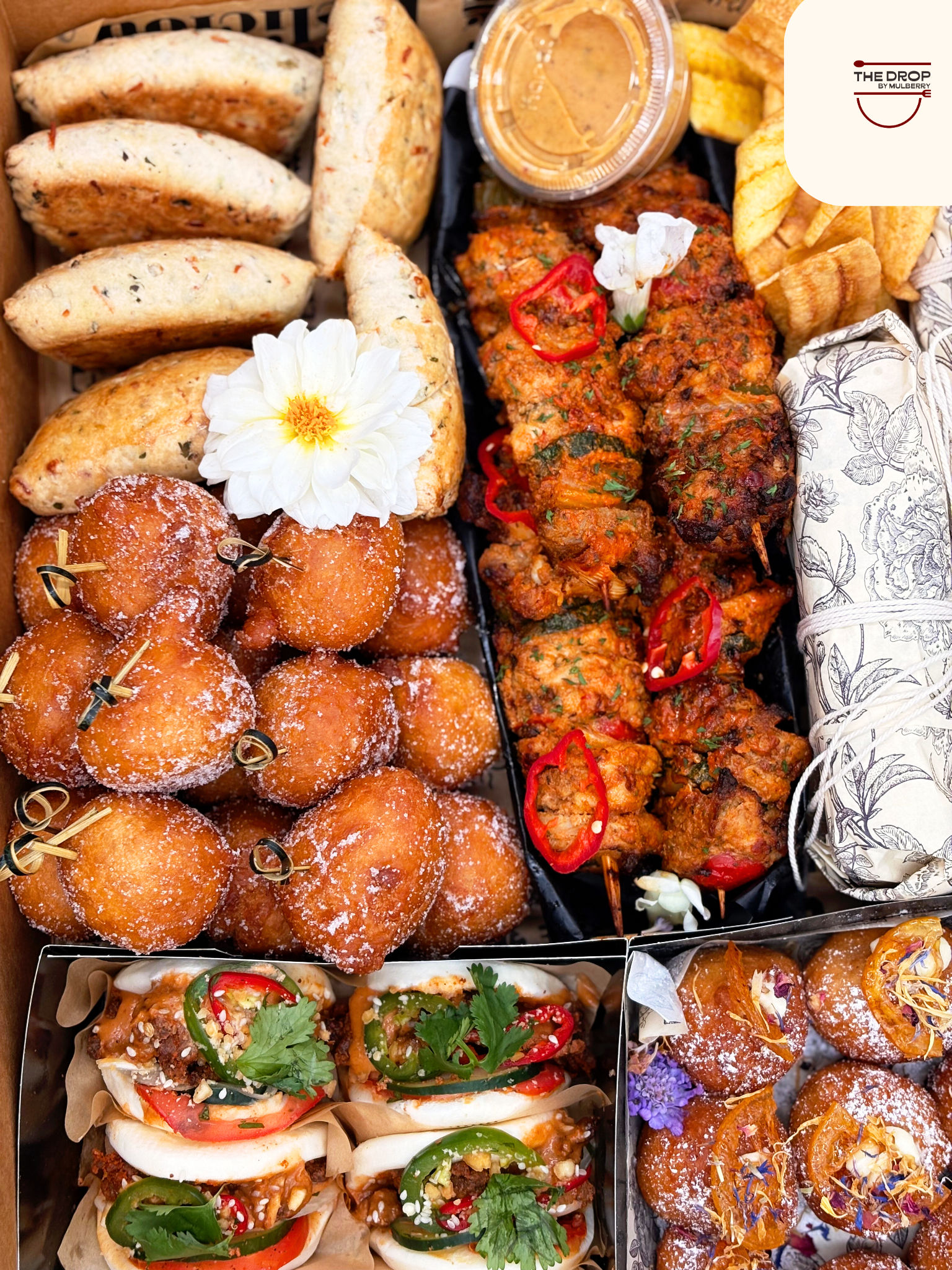 A tray of assorted Indian and Asian appetizers including samosas, fried cheese balls, kebabs, and stuffed buns, garnished with herbs and flowers.