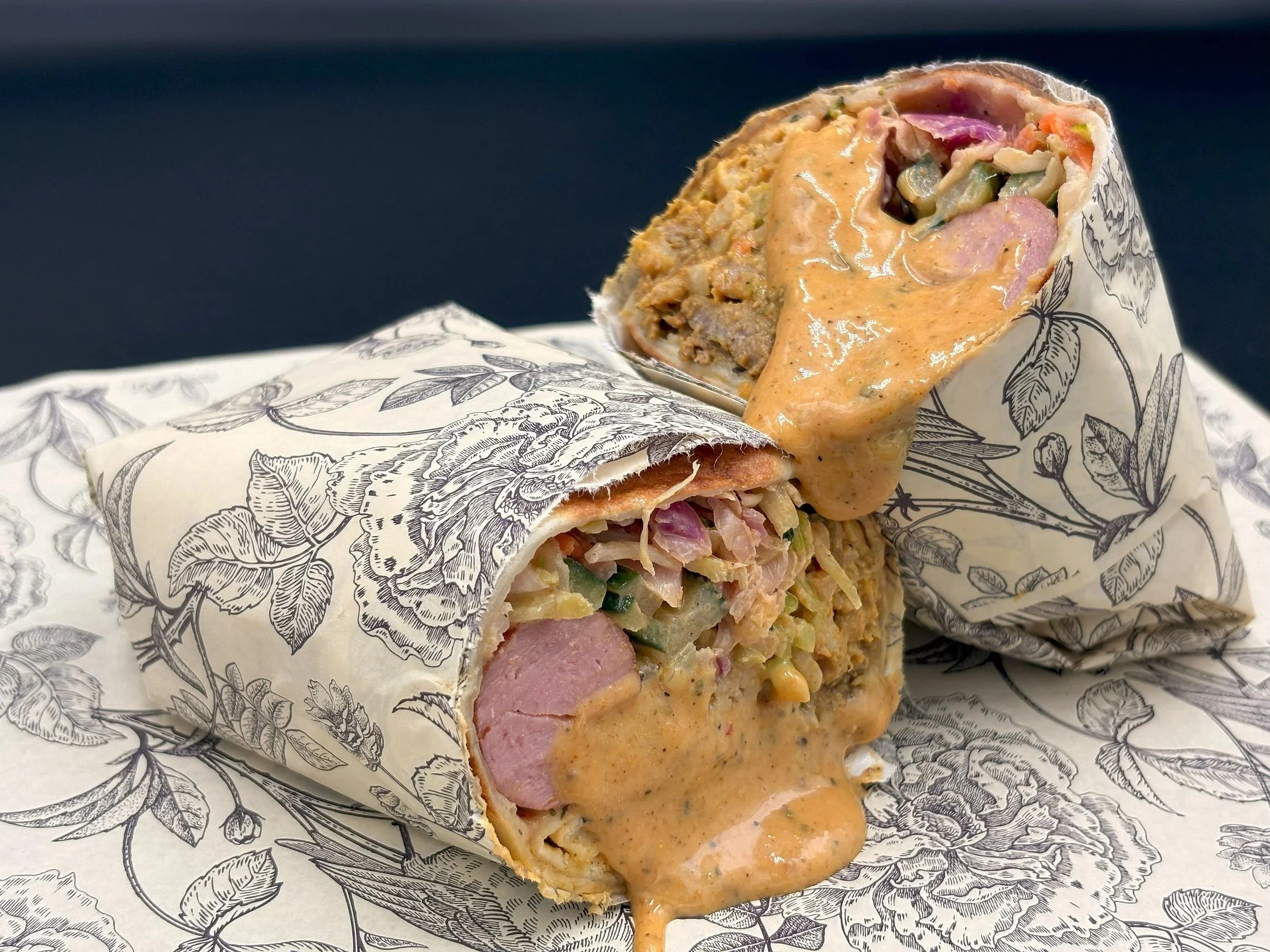 Two breakfast burritos with sliced sausage, scrambled eggs, chopped vegetables, and creamy sauce, wrapped in patterned paper.