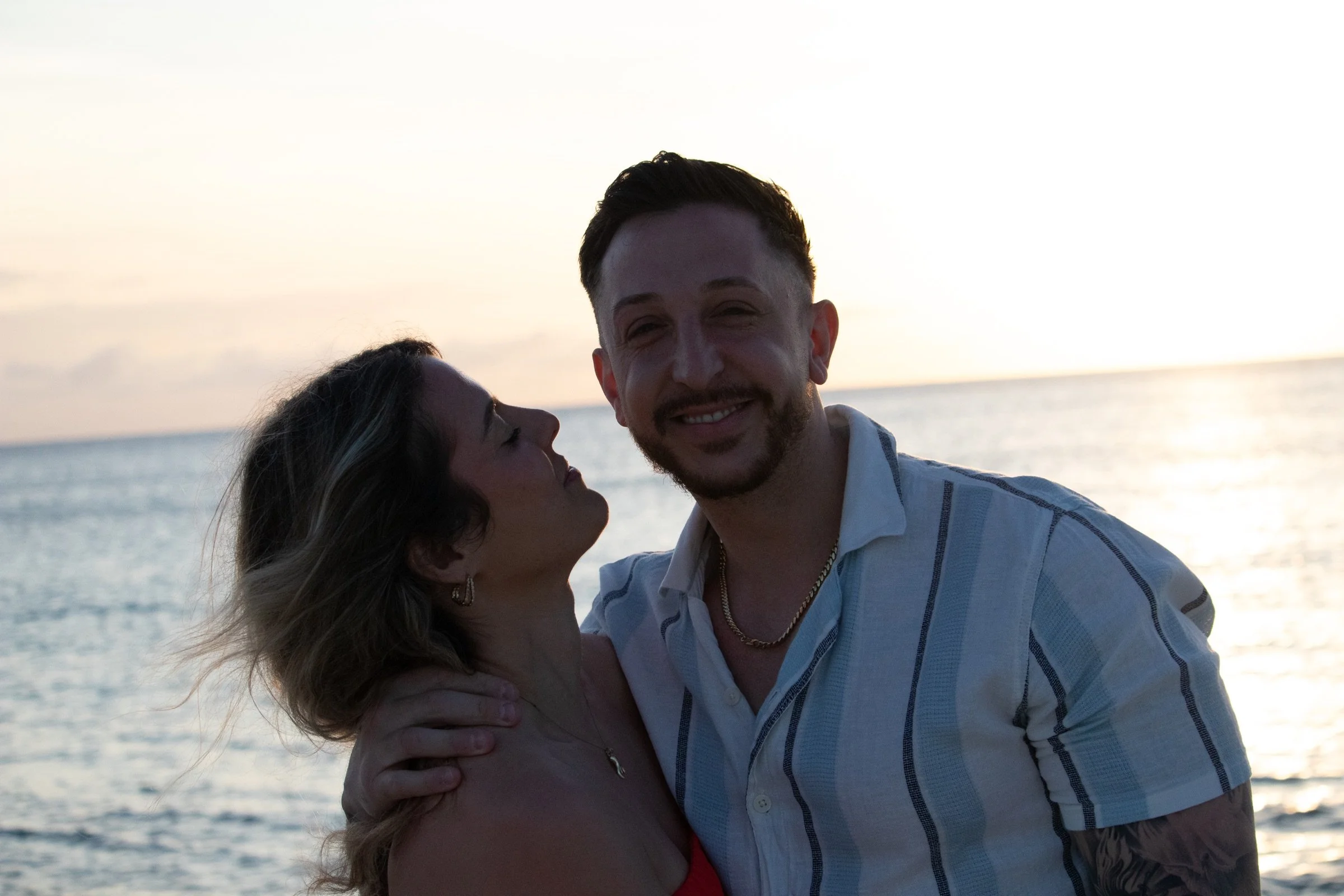 A newly engaged couple smiling together in warm light beside the Caribbean Sea.