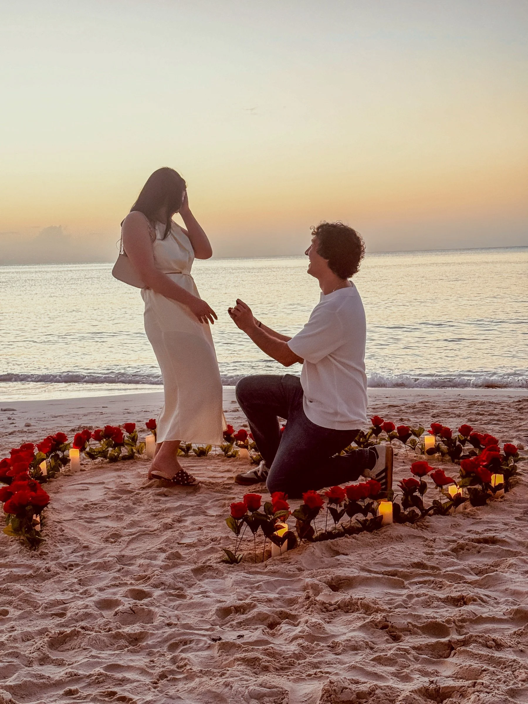 A romantic kneeling proposal inside a heart of roses and candlelight on the beach.