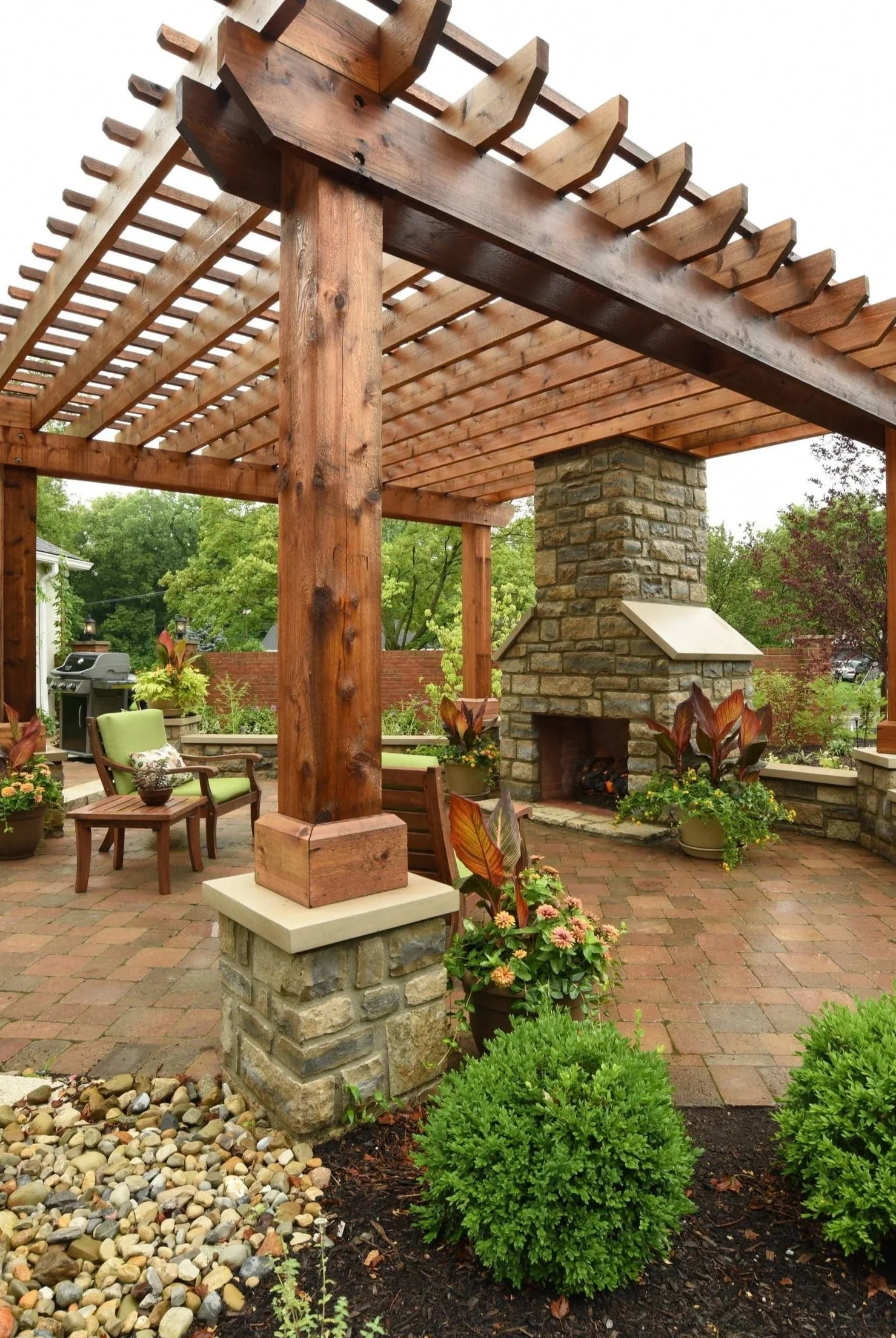 A backyard patio with a wooden pergola, stone fireplace, outdoor chairs, potted plants, and landscaped greenery.