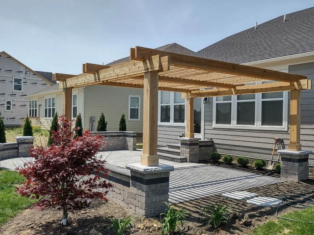 A newly constructed wooden pergola with brick pillars sits on a concrete patio in a backyard, with gardening plants and a house in the background.