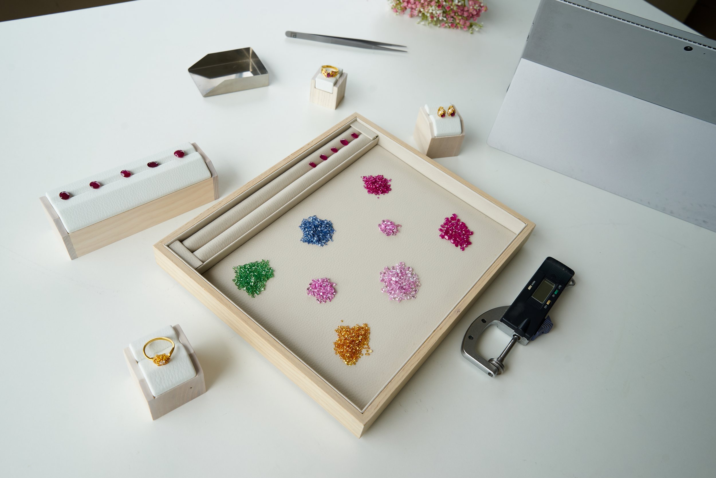 Jewelry design workspace with colorful gemstones, jewelry tools, and display cases on a white table.