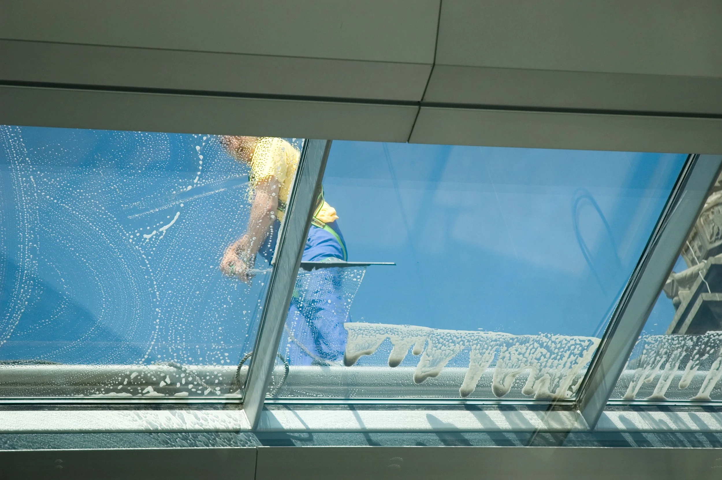 A window cleaner in yellow shirt and blue pants cleaning a large glass window on the roof of a building, with soap suds and water on the glass, against a clear blue sky.