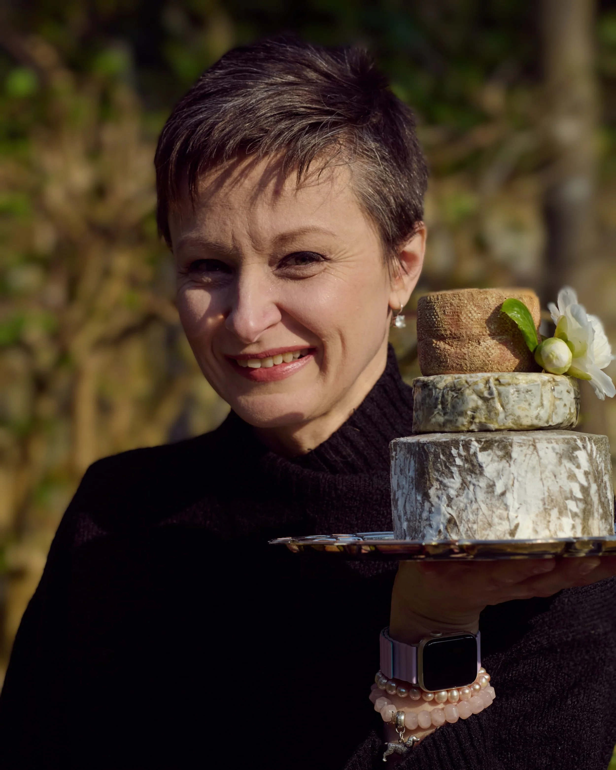 A woman with short brown hair smiling while holding a three-tiered cake with marble and brick-like textures, decorated with a white flower and green leaves outdoor in sunlight.