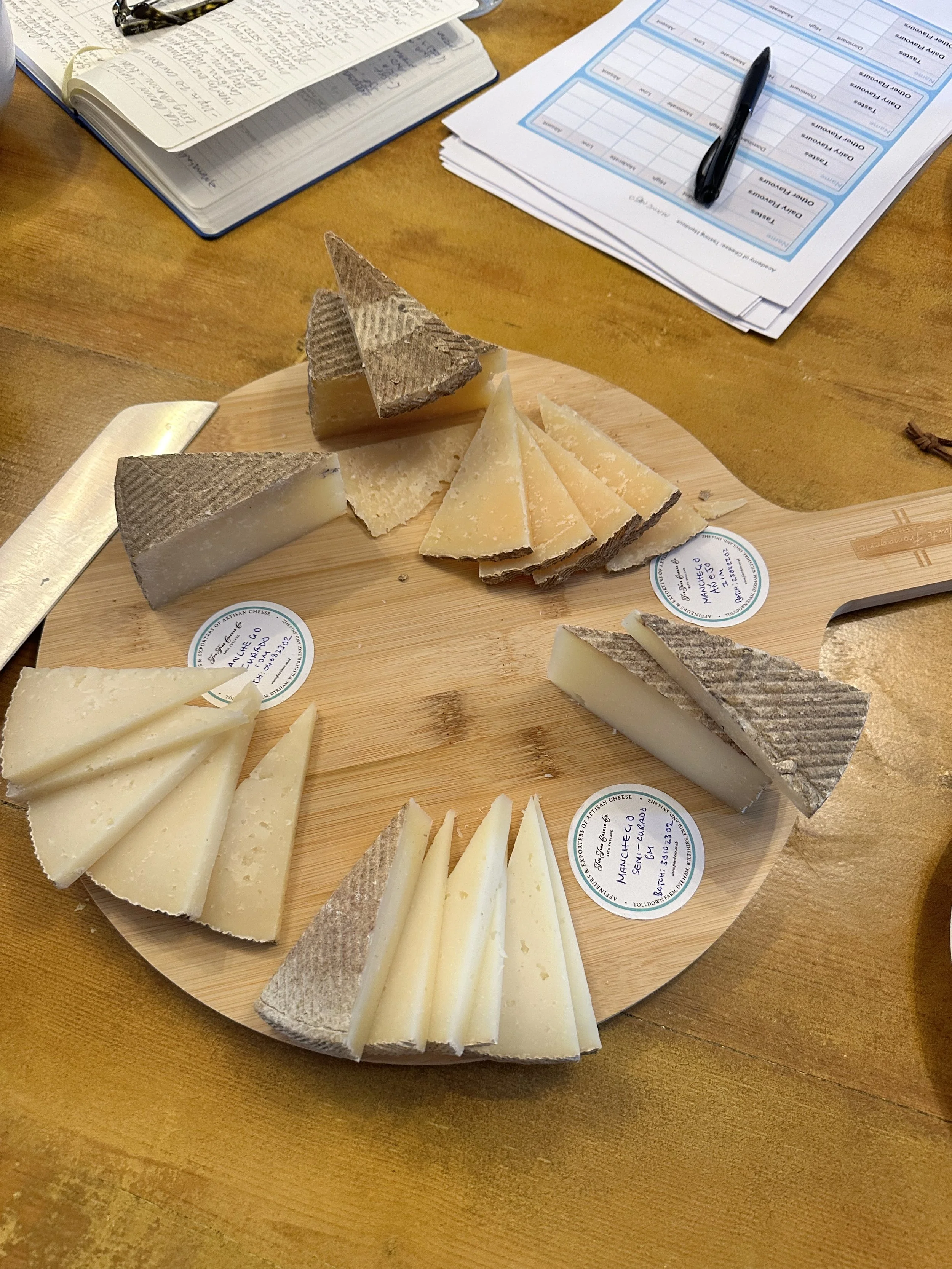 Platou de branzeturi rotund, de lemn, cu branzeturi taiate tringhiular langa fise de degustare si caiete de notite pentru cursul avansat de branzeturi