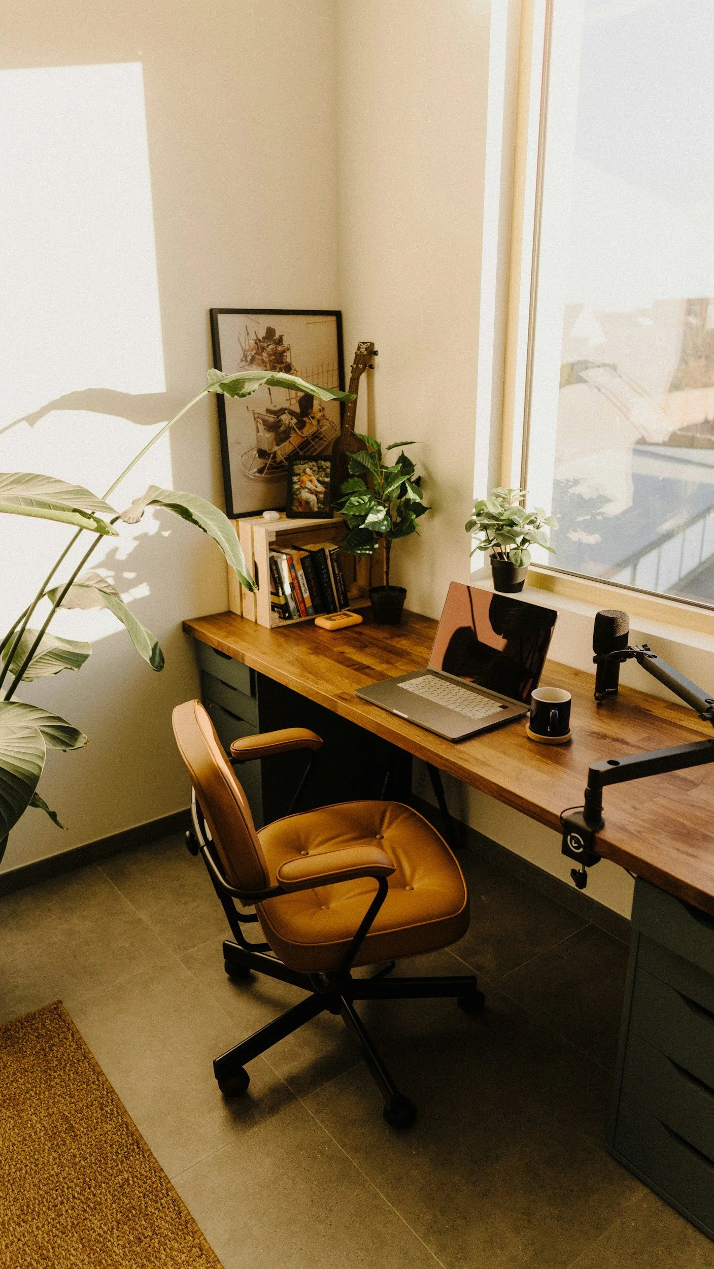 A cozy home office with a wooden desk by a large window, a brown office chair, a laptop, a coffee mug, a microphone, various potted plants, books, and a framed picture, all illuminated by natural light.