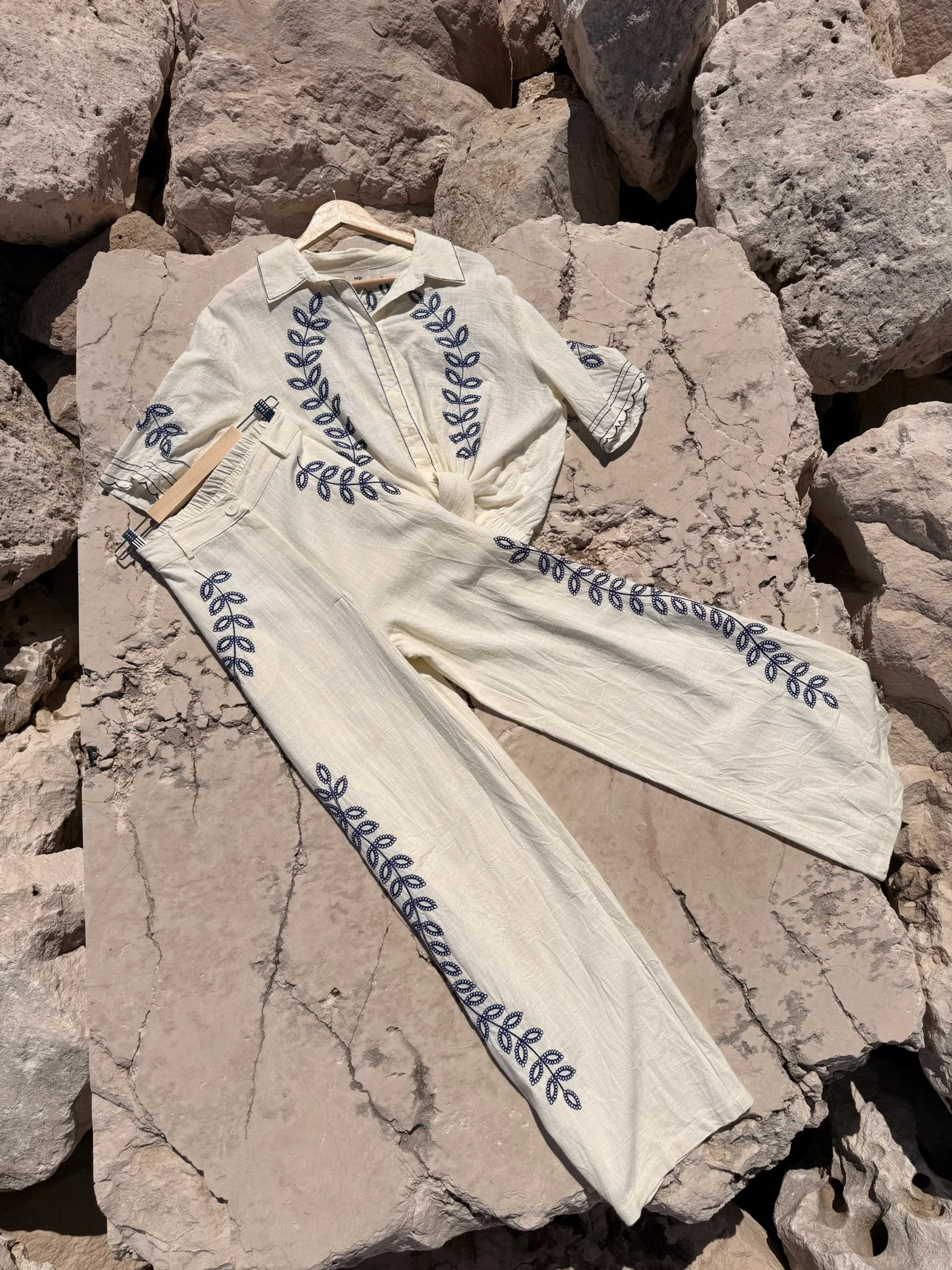 A cream-colored linen shirt and pants set with blue embroidered leaf patterns, laid on a rocky surface.