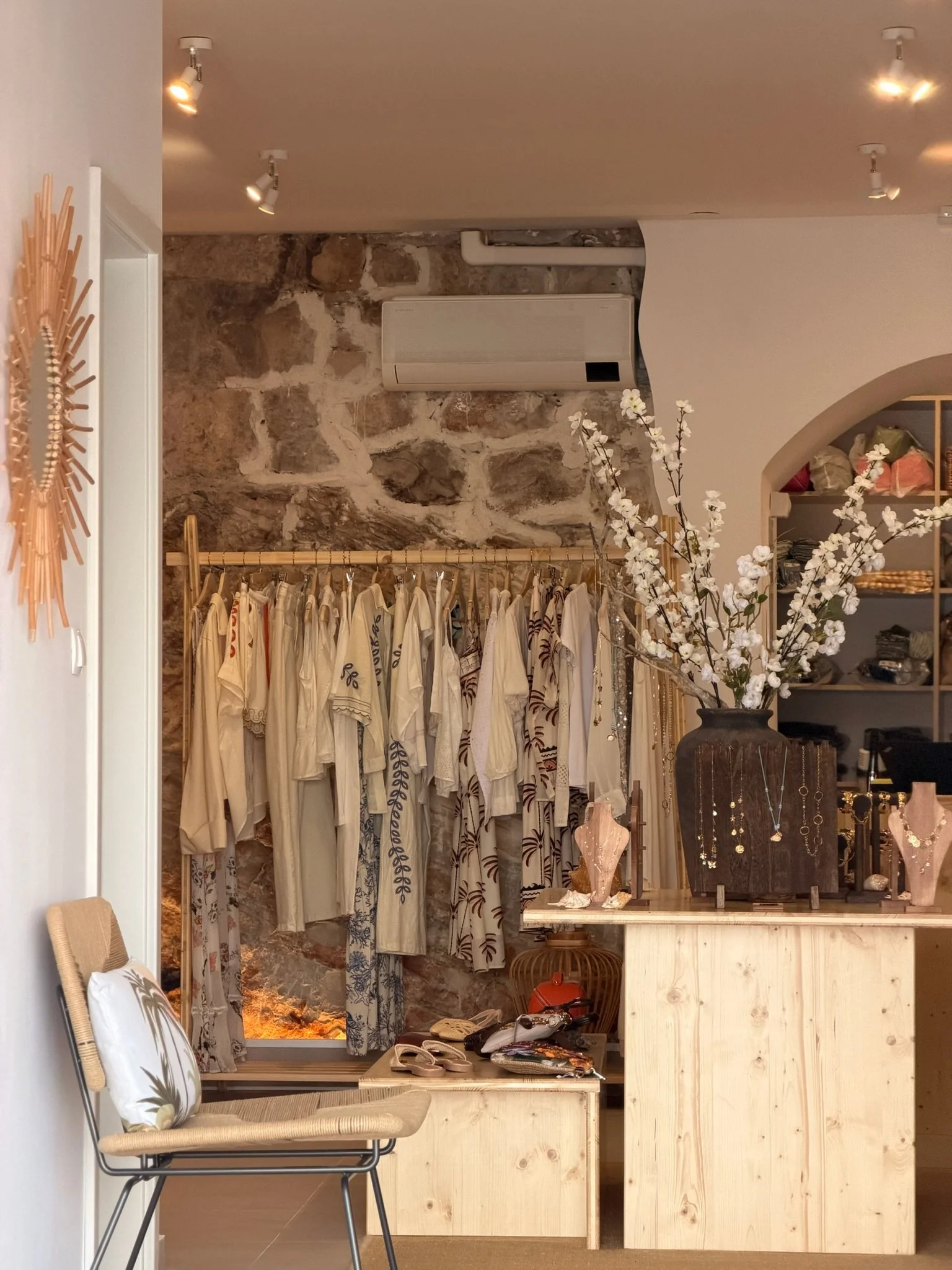 Display of clothing on a rack with dresses, a large flower vase with white flowers on the counter, and jewelry on display. A seating chair with a decorative pillow is in the foreground. The background features a stone wall, air conditioning unit, and ceiling spotlights.