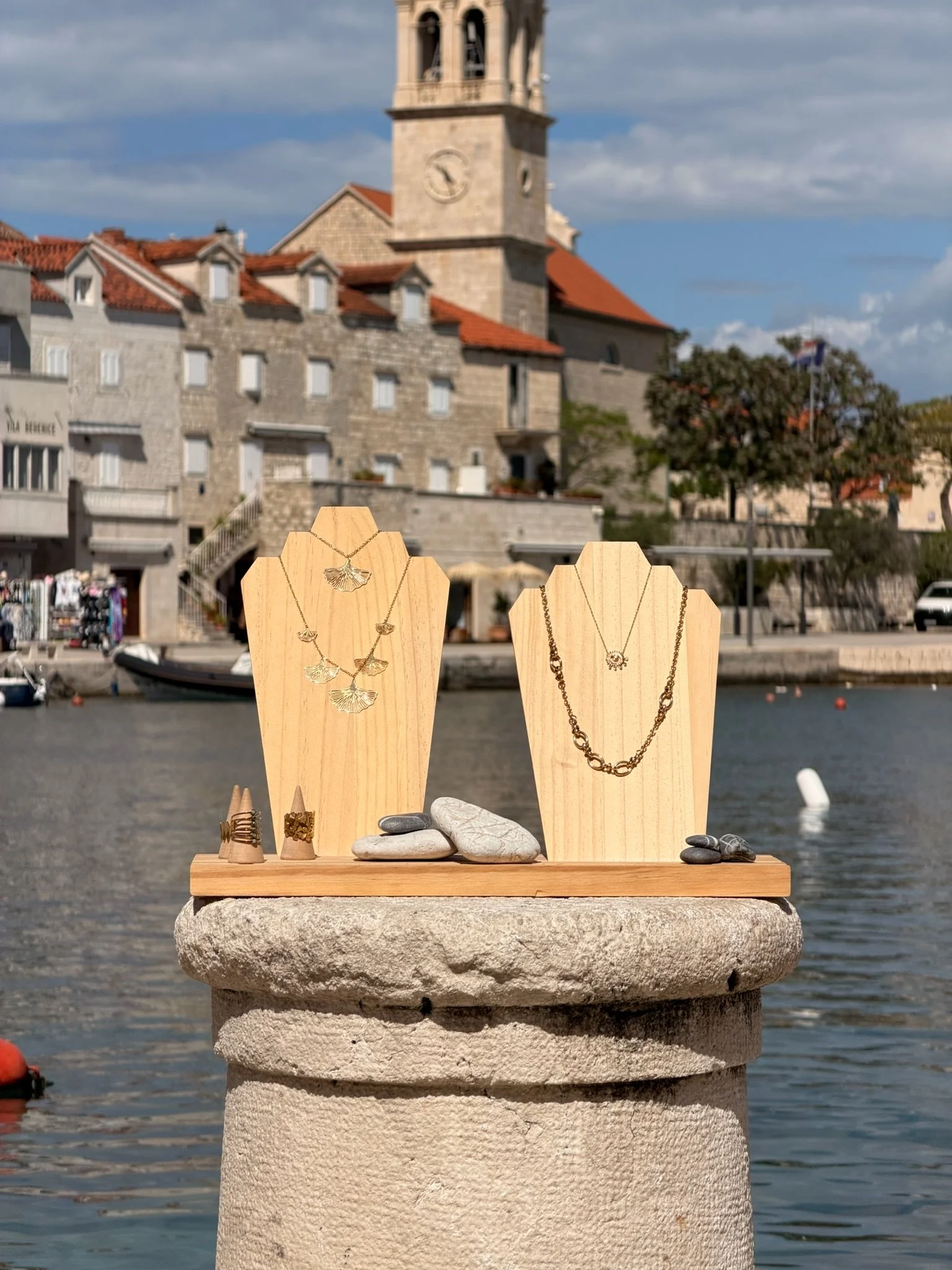 Jewelry display with necklaces and rings on wooden stands, set on a stone pedestal near a waterfront with historic buildings and boats in the background.