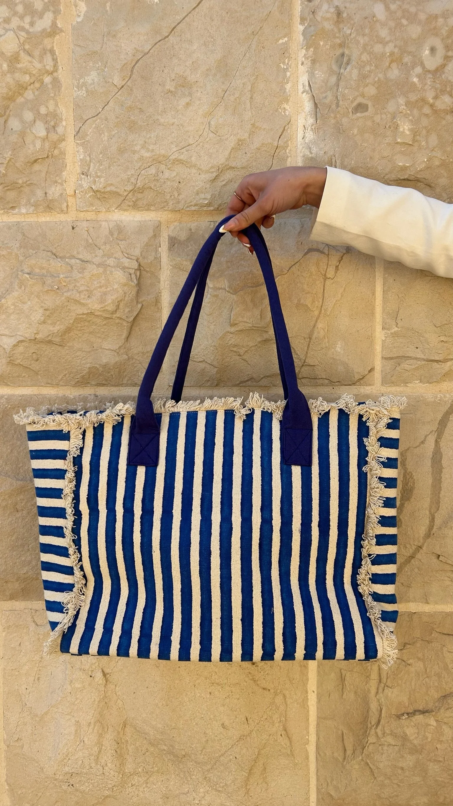 A hand holding a blue and white striped tote bag with fringed edges against a beige stone wall.