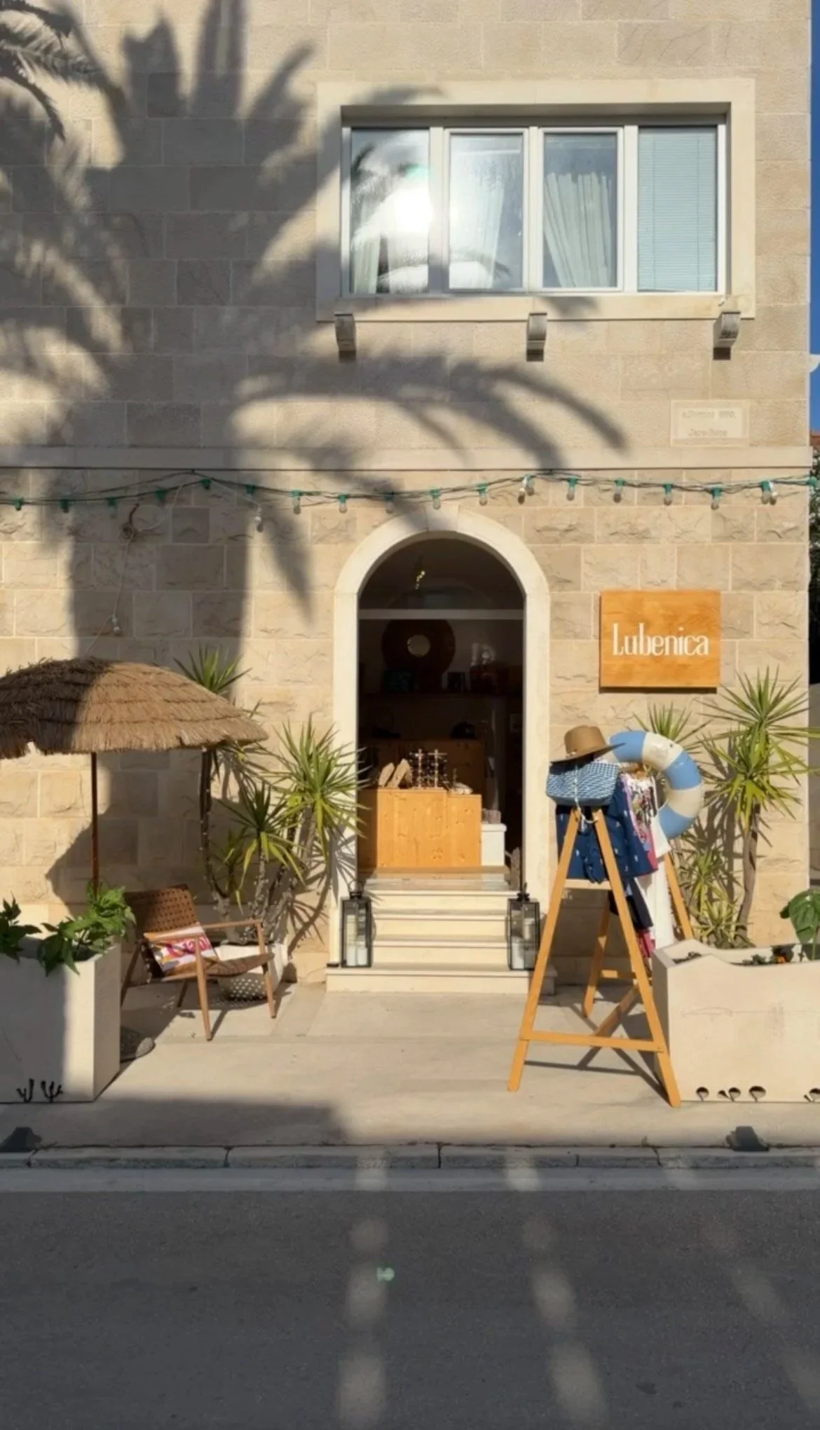 Storefront with a sign that says 'Lubenica,' surrounded by potted plants, outdoor seating, a thatched umbrella, and decorative items.