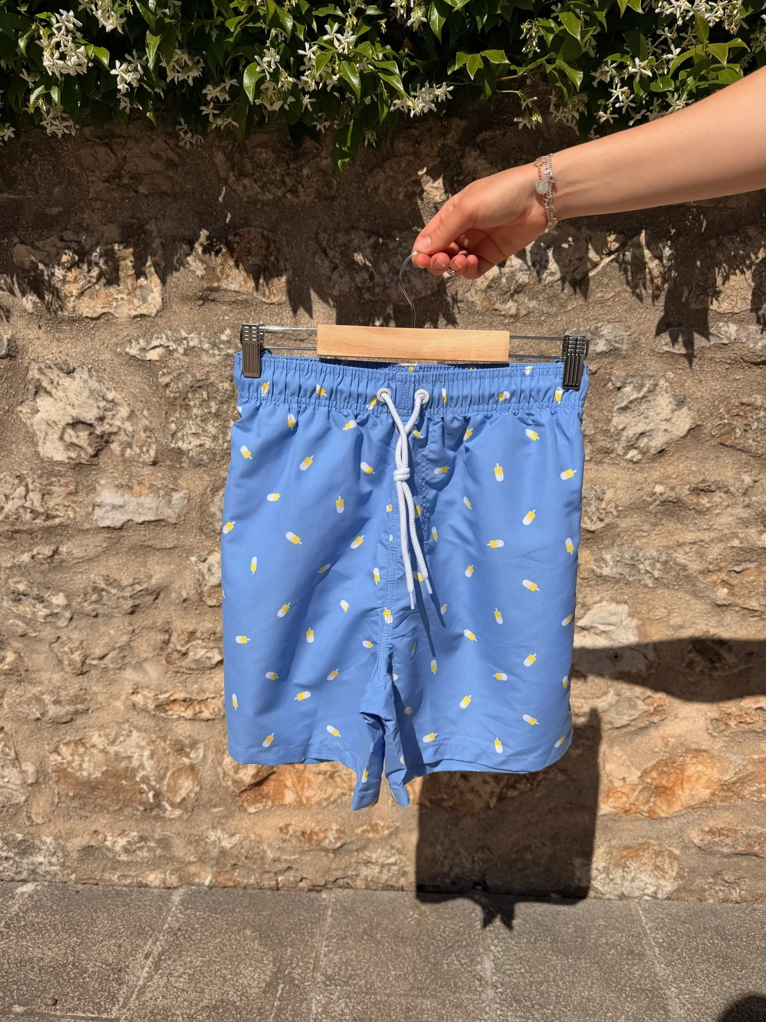 Blue swim shorts with a white drawstring, hanging on a hanger, against a stone wall with greenery above.