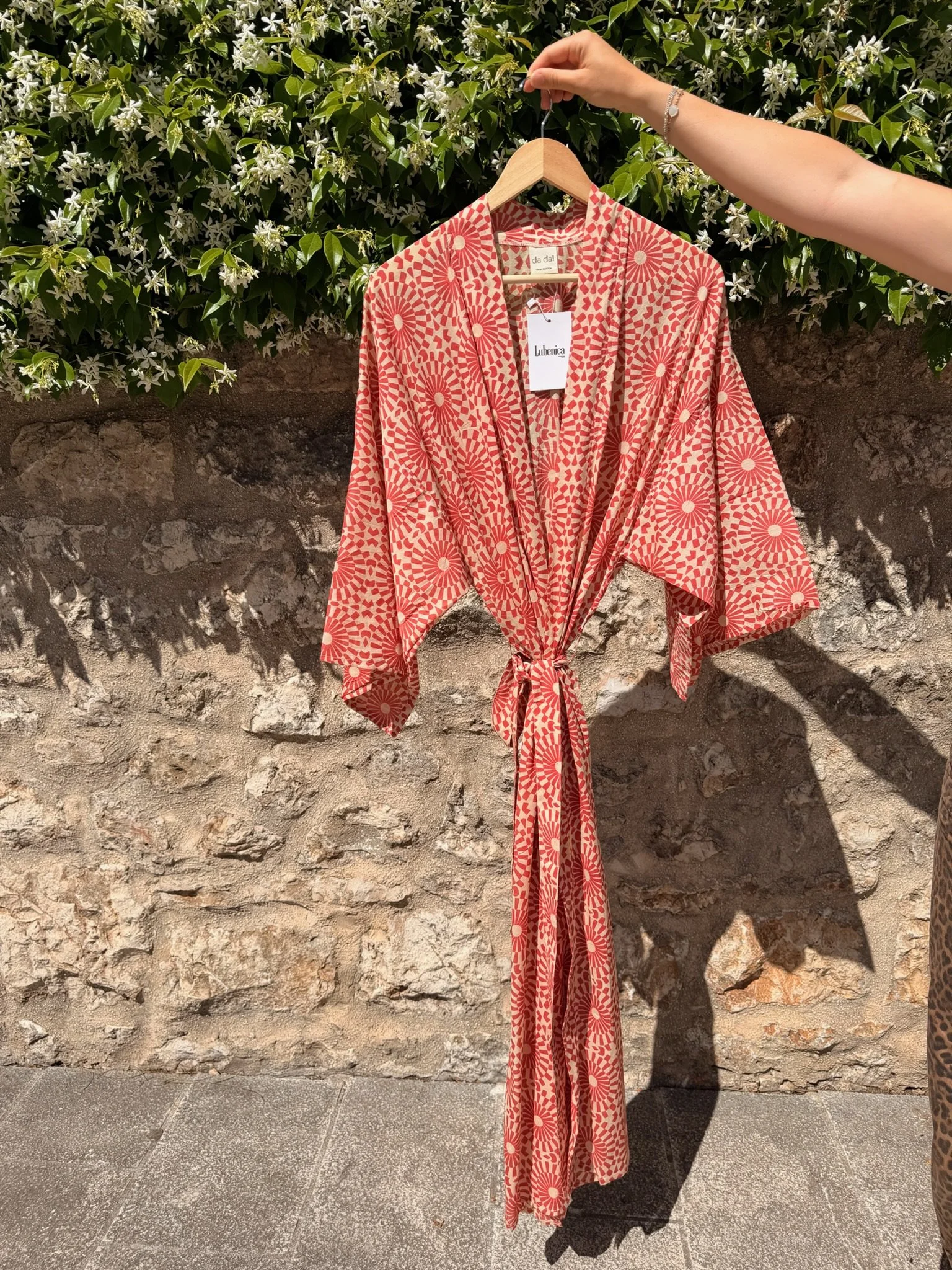 A peach-colored kimono with a red and white geometric pattern hanging on a wooden hanger against a stone wall and green foliage with white flowers.