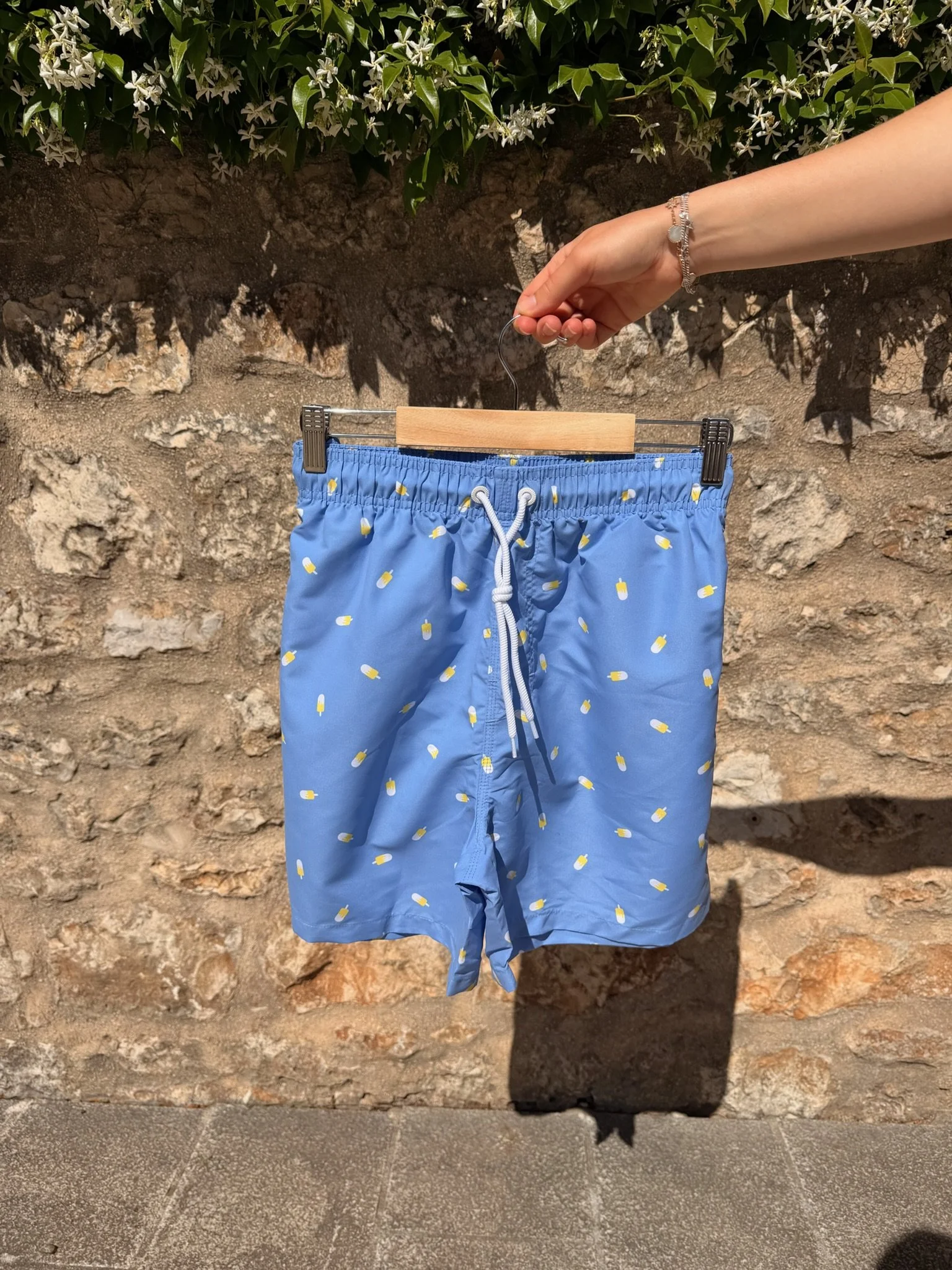 Blue patterned swimming shorts hanging on a wooden hanger with a person's hand, wearing a silver bracelet, holding the hanger, against a stone and plant background.