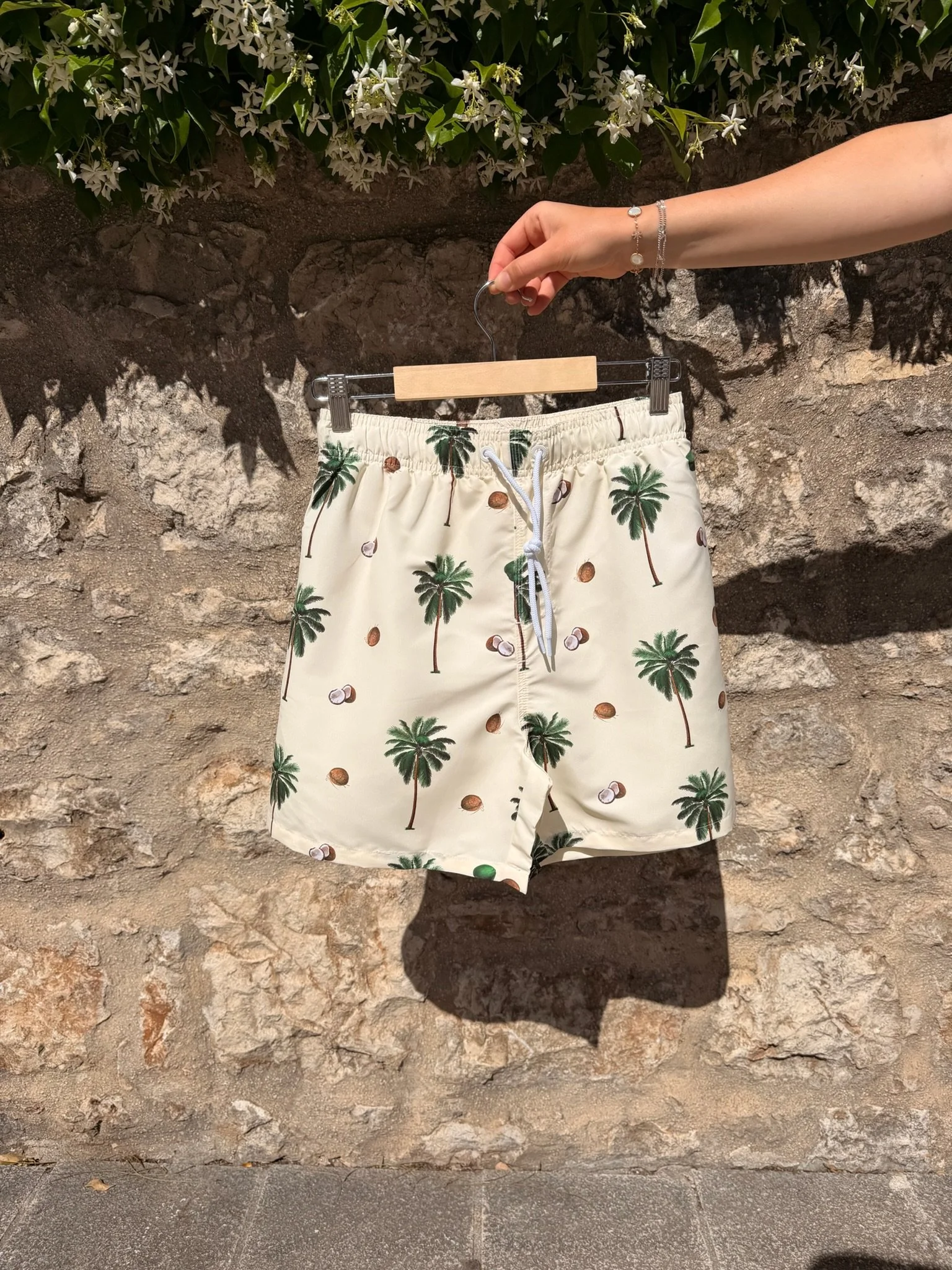 A pair of beige shorts with palm tree and coconut prints hanging on a hanger against a stone wall. The shorts have a white drawstring and are adorned with jewelry on the person's wrist.