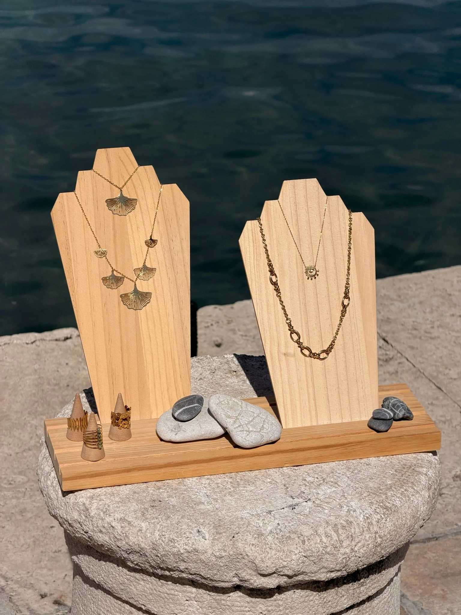 Jewelry display featuring necklaces, earrings, rings, and stones on wooden stands and a stone surface, outdoors near water.