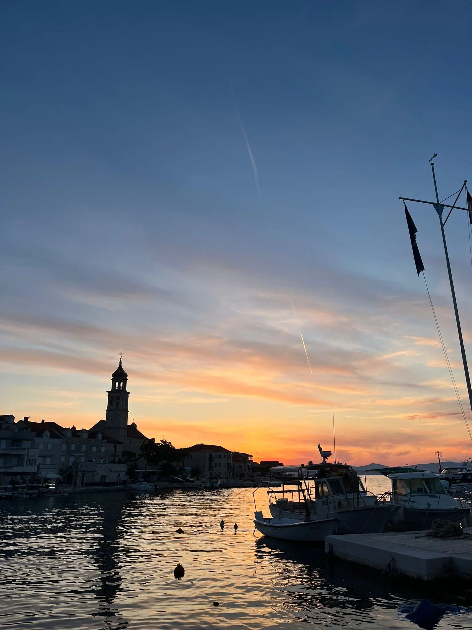A view of a harbor at sunset with boats moored along the pier, a church steeple, and a flagpole with flags. The sky is painted with warm hues of orange, pink, and purple as the sun sets behind the buildings.