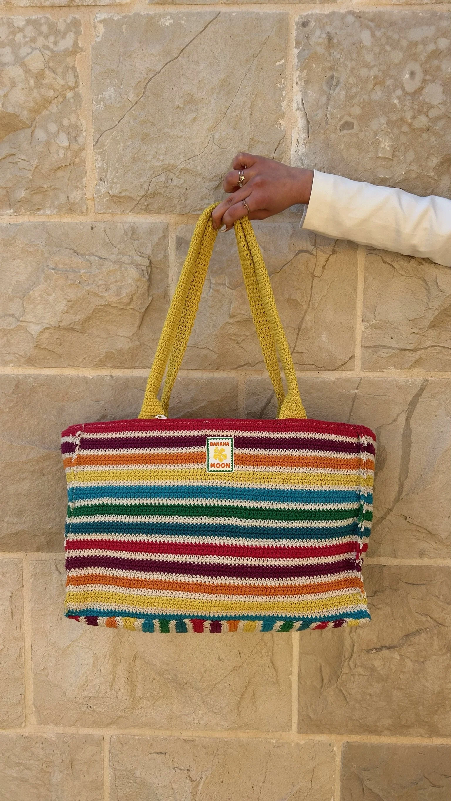 A colorful striped handbag hanging on a person's hand against a stone wall. The person is wearing a white sleeve and gold rings. The handbag has a label that reads 'Banana Moon' with a yellow banana logo.