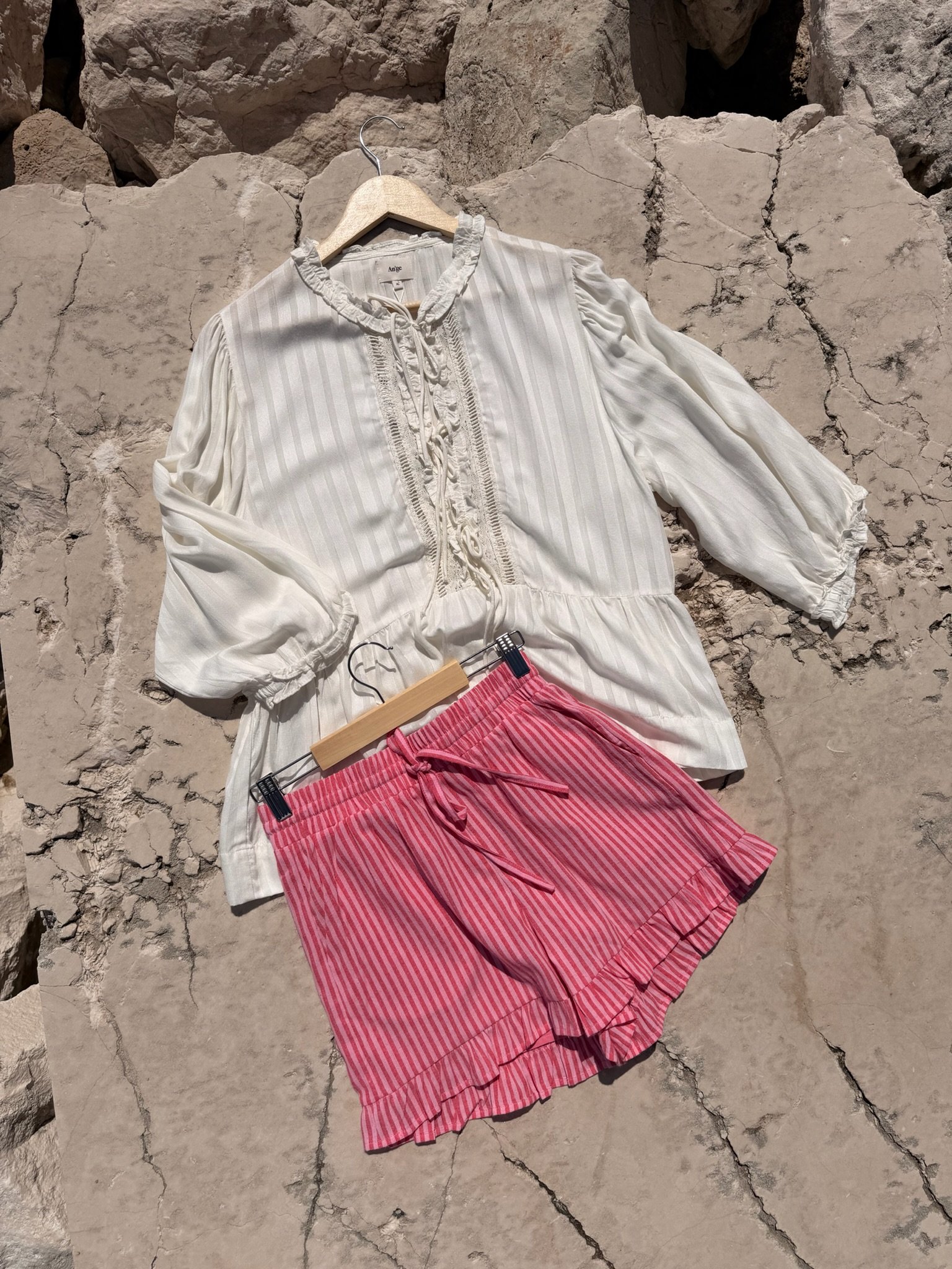 A white blouse hanging on a wooden hanger and a pink striped skirt hanging on a black clip, placed on a rough, cracked stone surface.