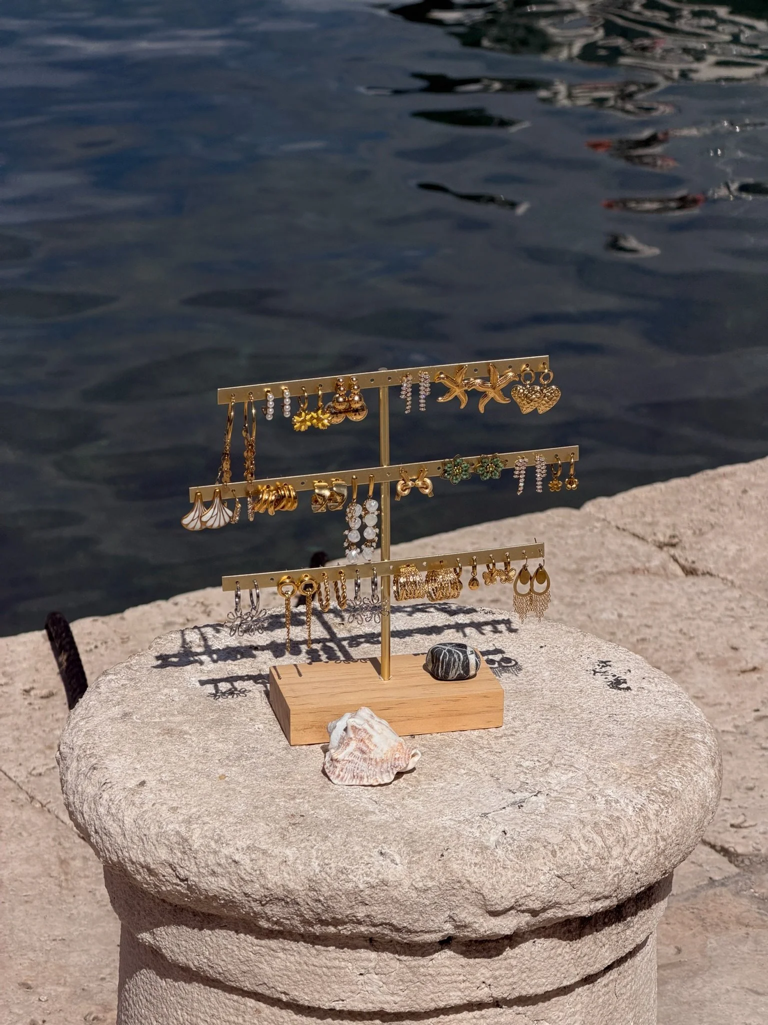 A jewelry display stand with earrings and necklaces, placed on a stone surface near water.
