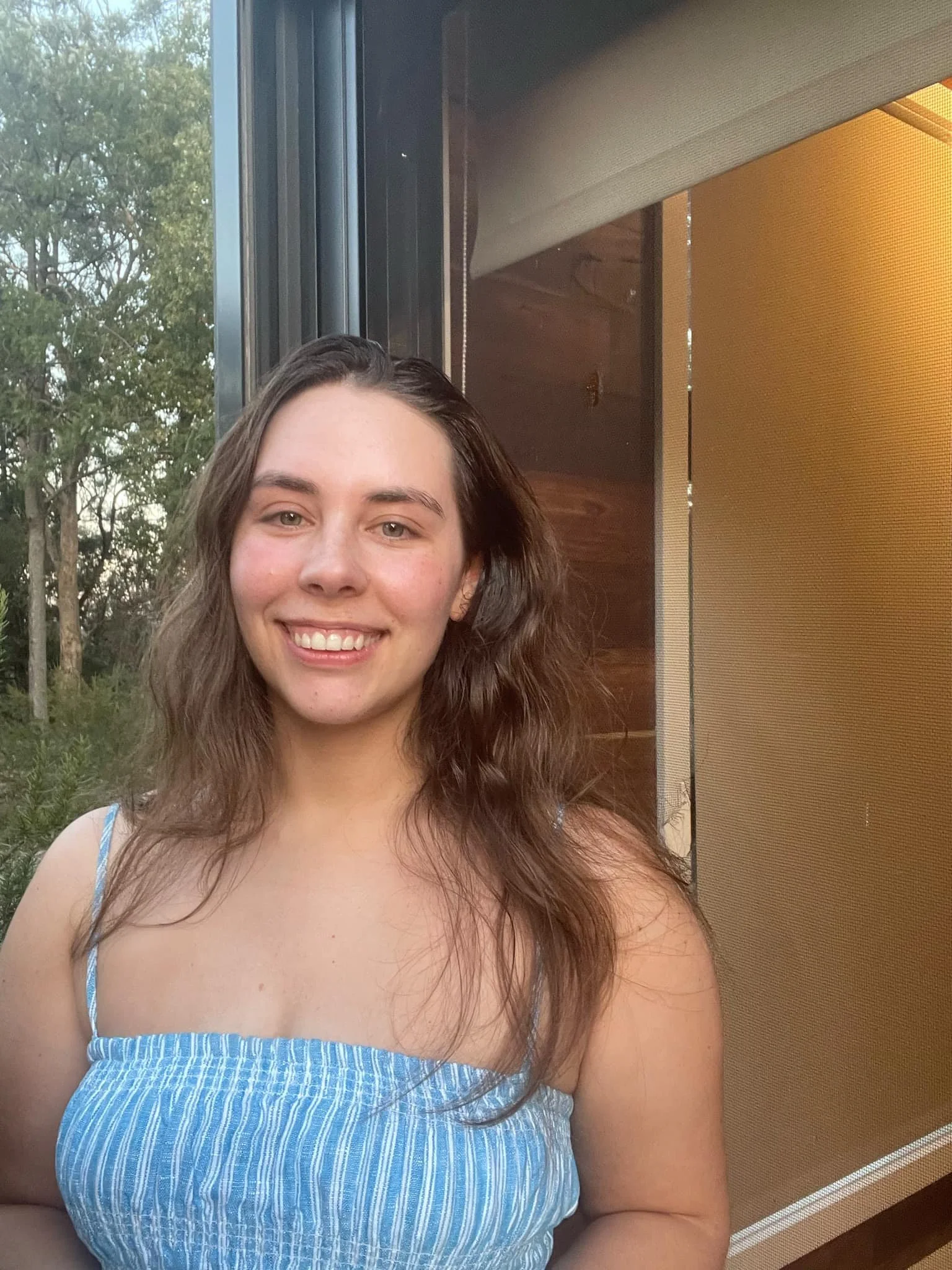 A young woman with long brown hair and fair skin smiling and wearing a blue and white striped sleeveless dress, standing in front of a window with trees visible outside and a wooden cabinet with a yellow panel to her right.