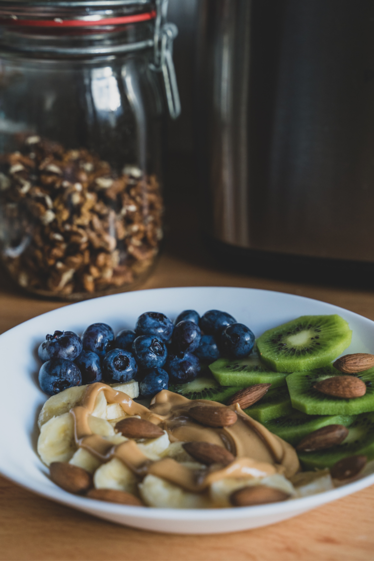A white bowl with blueberries, sliced kiwi, banana slices, almond nuts, and peanut butter. In the background, a jar of granola and a stainless steel appliance are visible.