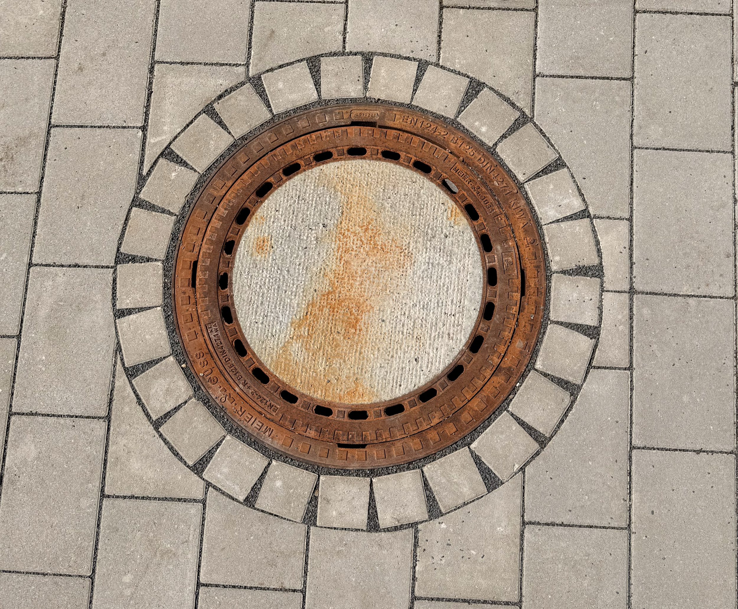 Aufgefundener runder Gullydeckel in der Gehwegpflasterung, mit rostbrauner Farbe und einem Muster aus schwarzen Steinen ringsum.