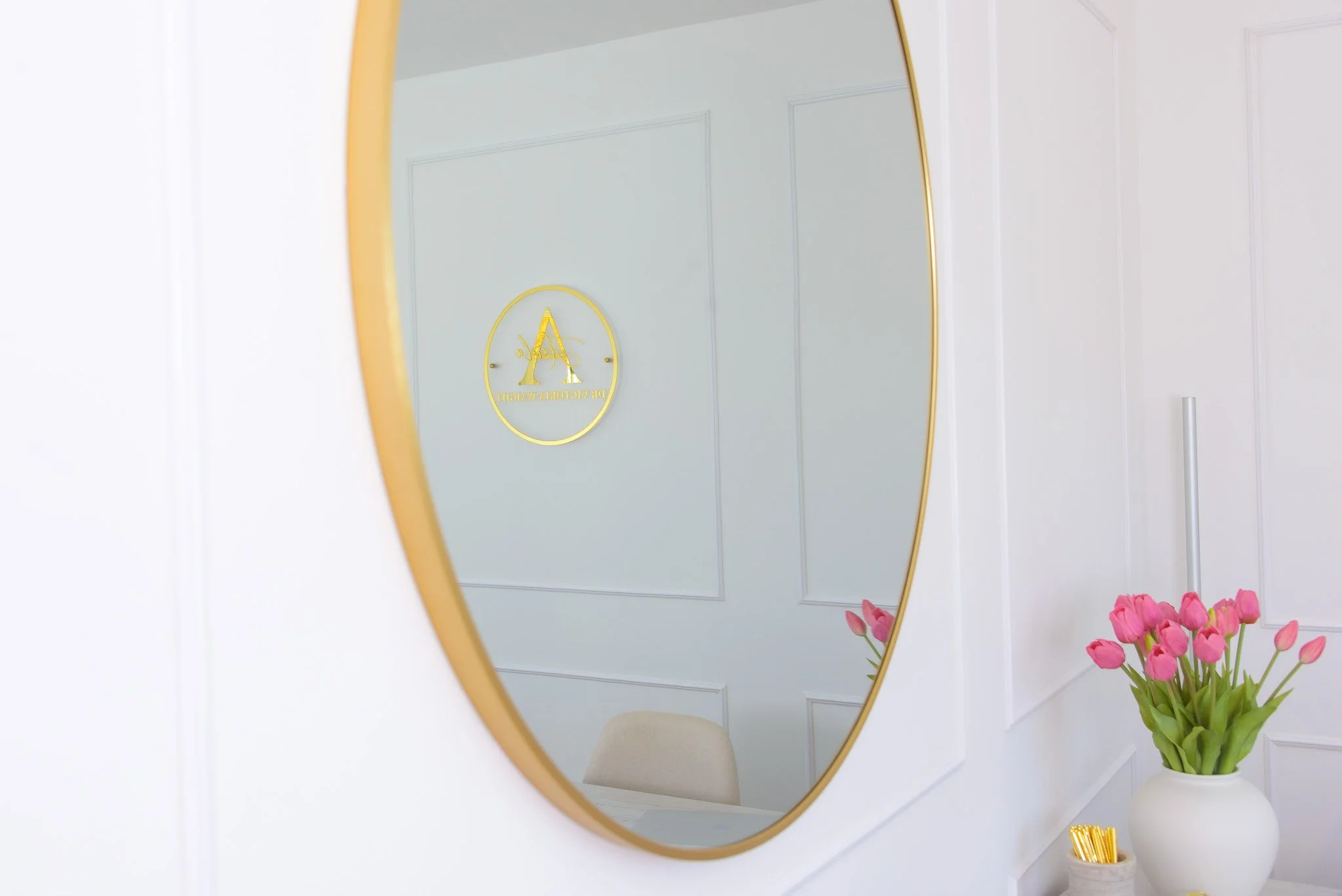 A mirror with a gold frame on a white wall, reflecting a logo on the wall that features a large gold letter 'A' within a circle. To the right of the mirror, there is a white vase filled with pink tulips on a small table or shelf.