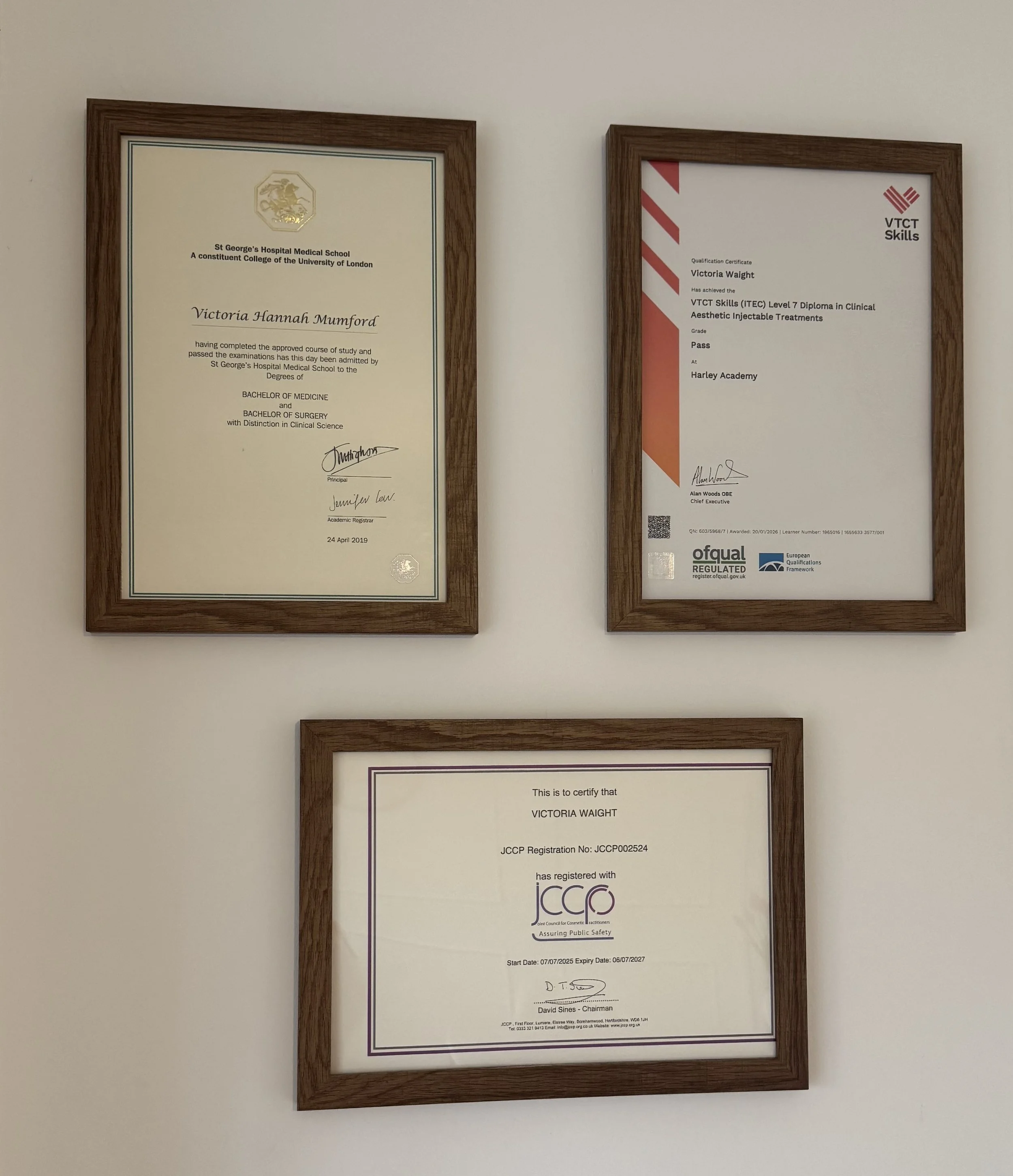 Three framed certificates hanging on a white wall, two on top and one below. The top left certificate is from St. George's Hospital Medical School, awarded to Victoria Hannah Mumford. The top right certificate is from VTCT Skills, awarded to Victoria