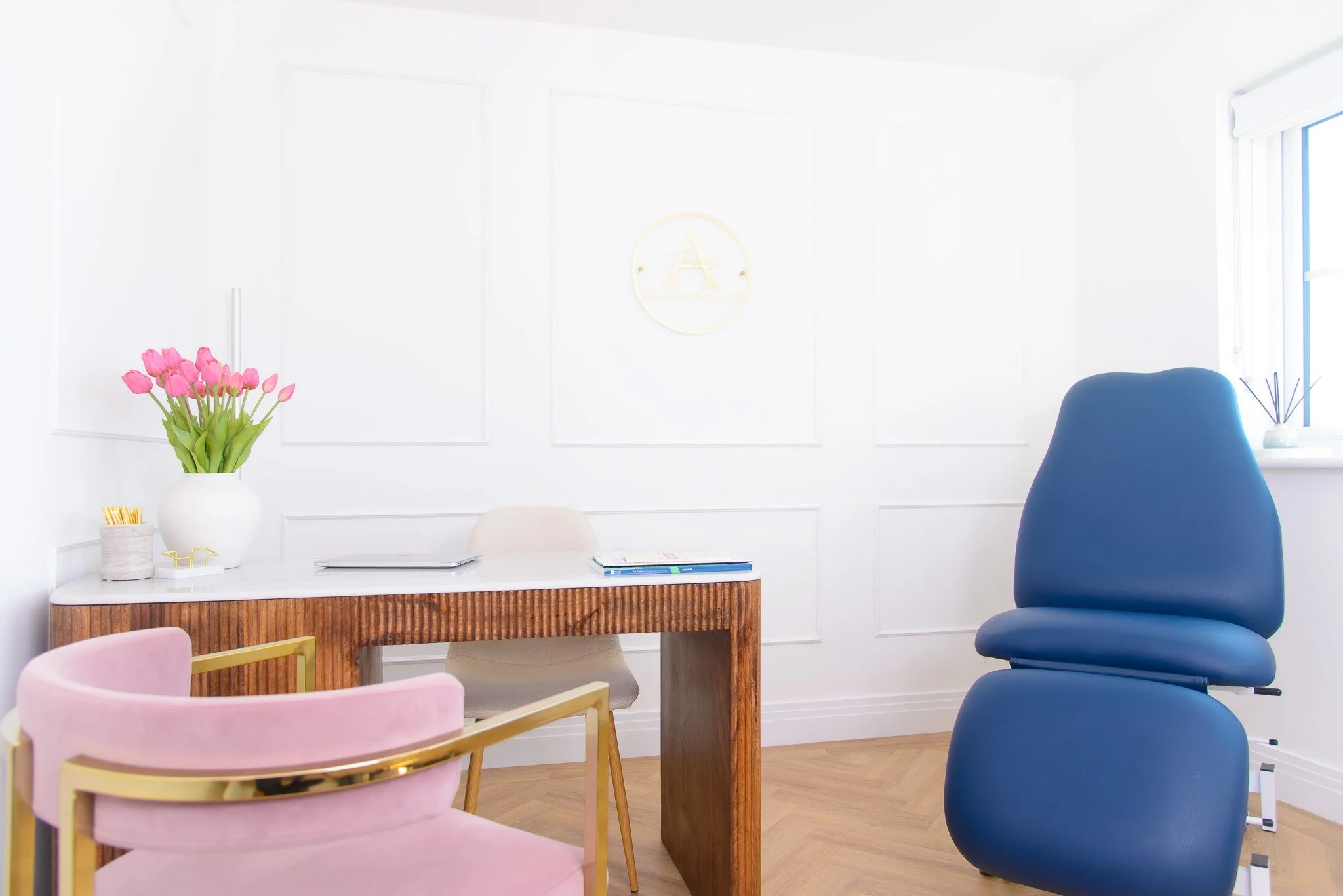 Clinic room of Aurela Aesthetics in Chichester. Bright modern office with a white wall, pink vase of tulips, wooden desk, blue ergonomic chair, pink chair with gold frame, and a window with blinds.