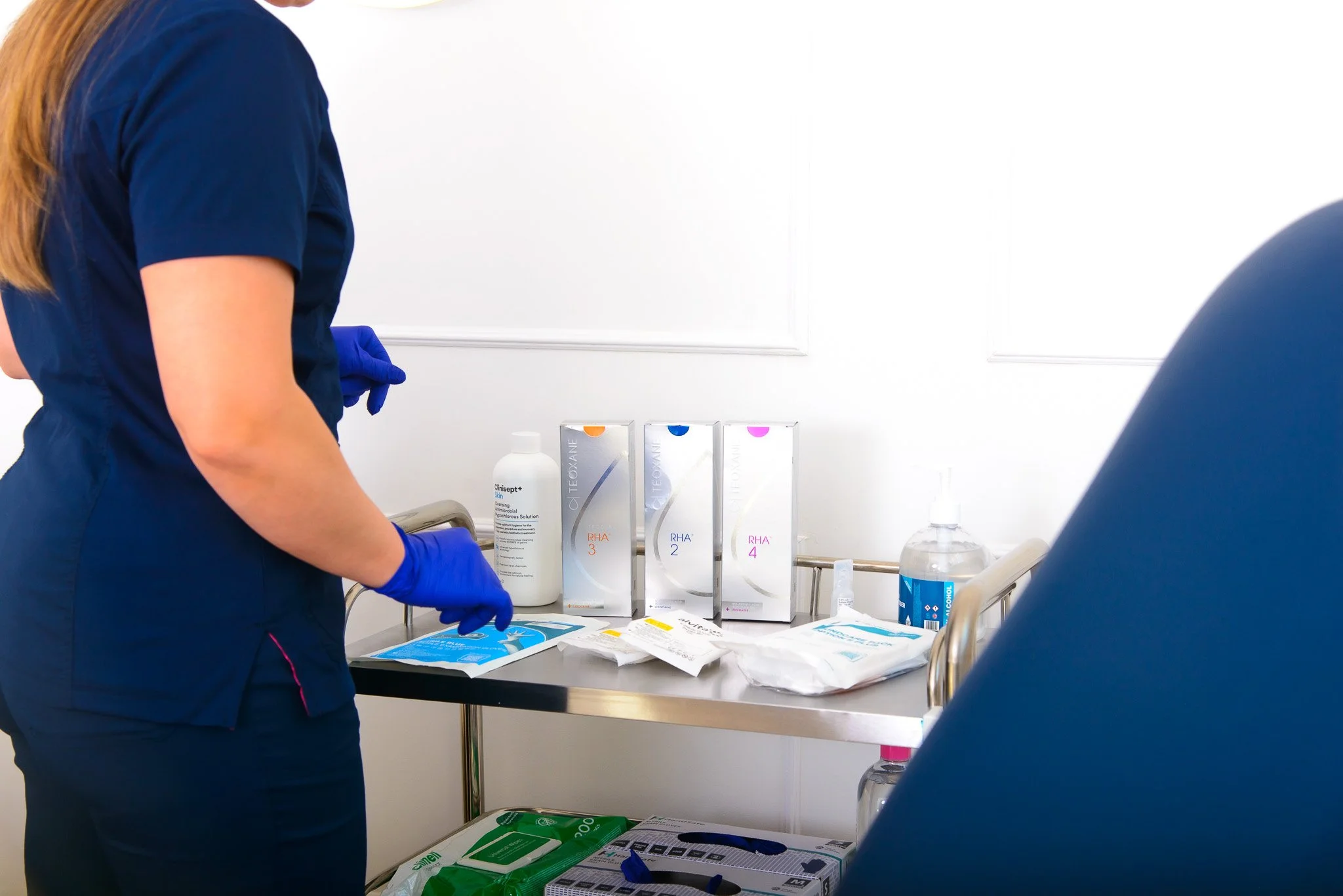 Medical professional wearing navy scrubs and blue gloves working near a stainless steel medical cart with various bottles, boxes, and supplies, including hand sanitizer and packaging, in a clinical setting.