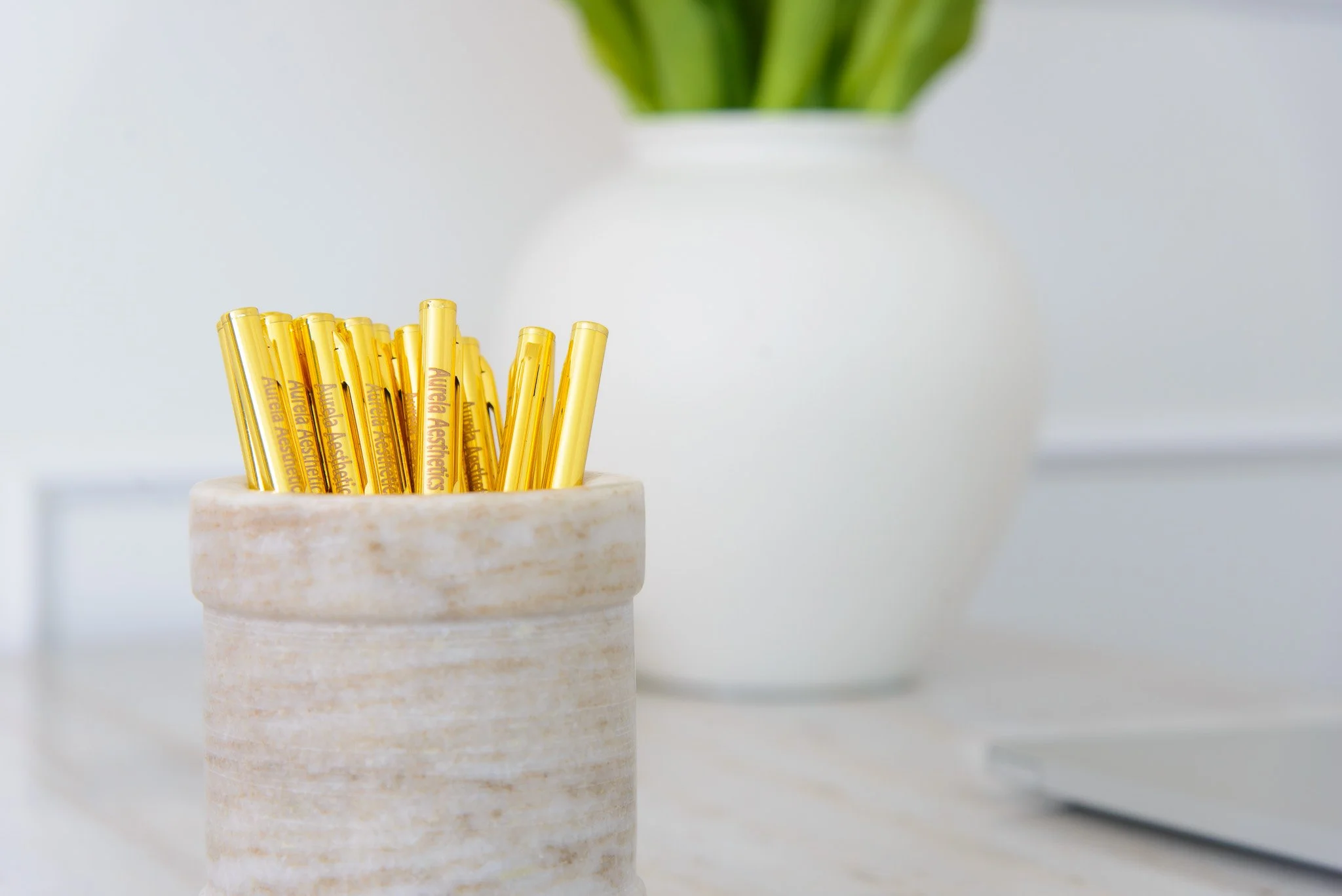 A container with yellow pens labeled 'Aurela Aesthetics' on a white surface, with a white vase holding green leaves in the background.
