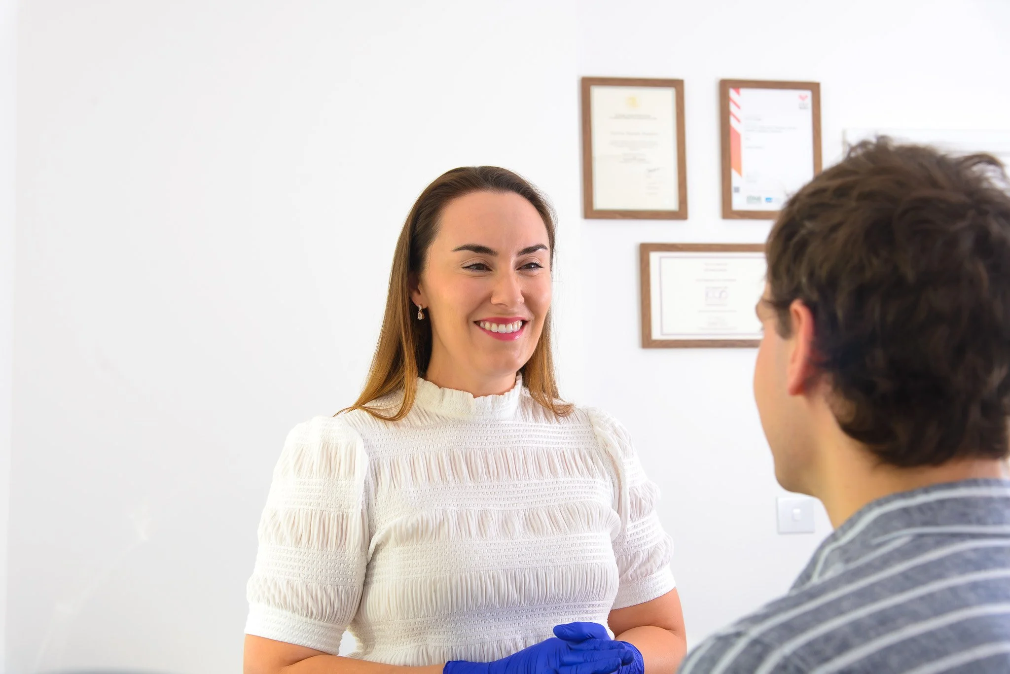 A smiling woman, Dr Victoria Waight, wearing a white top and blue gloves talking to a man with brown hair in a striped shirt in a clinical setting with framed certificates on the wall.