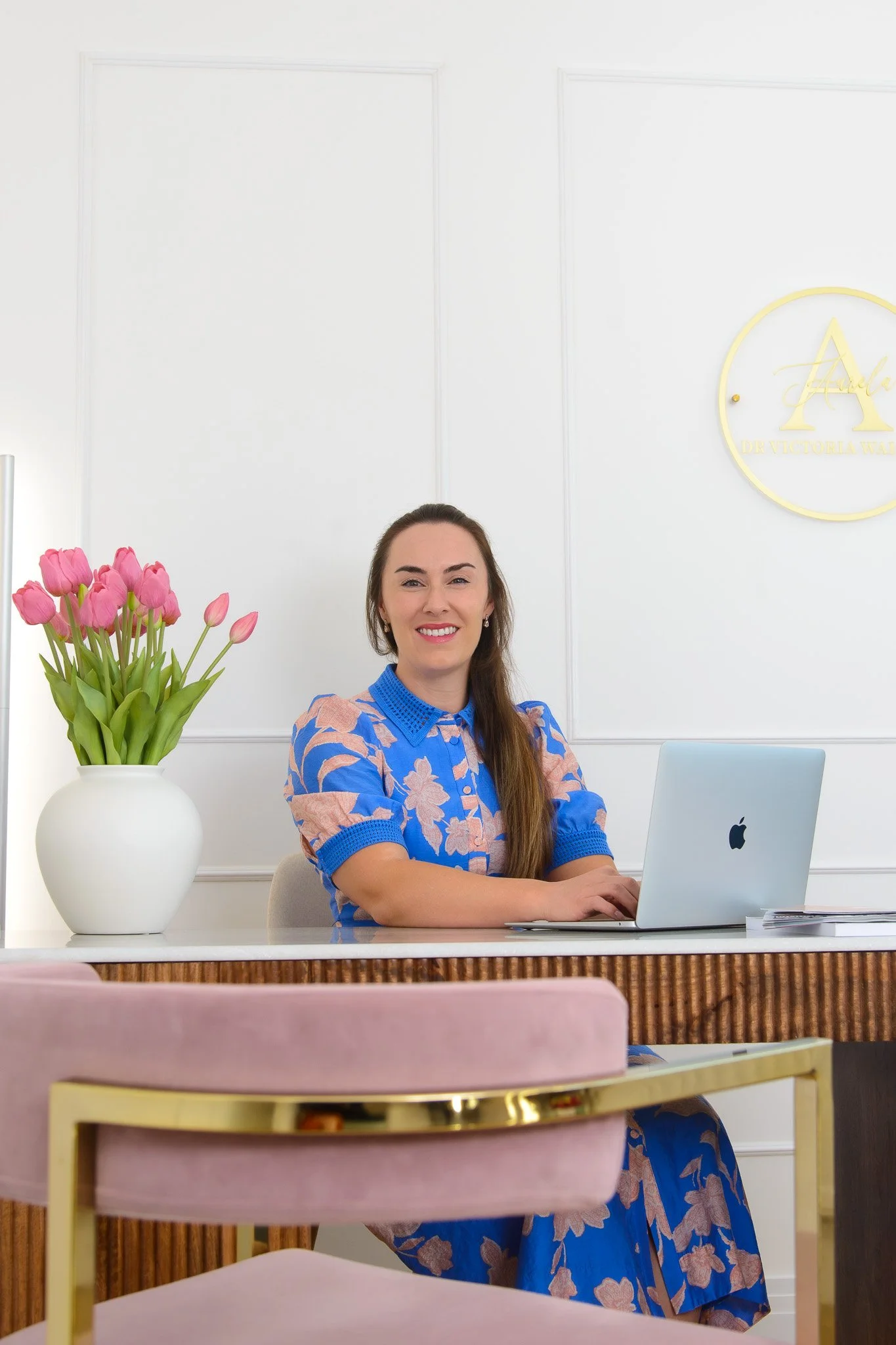 Dr Victoria Waight sitting at a desk with a silver laptop, smiling at the camera, with a white vase of pink tulips on her left in a bright, modern office space.