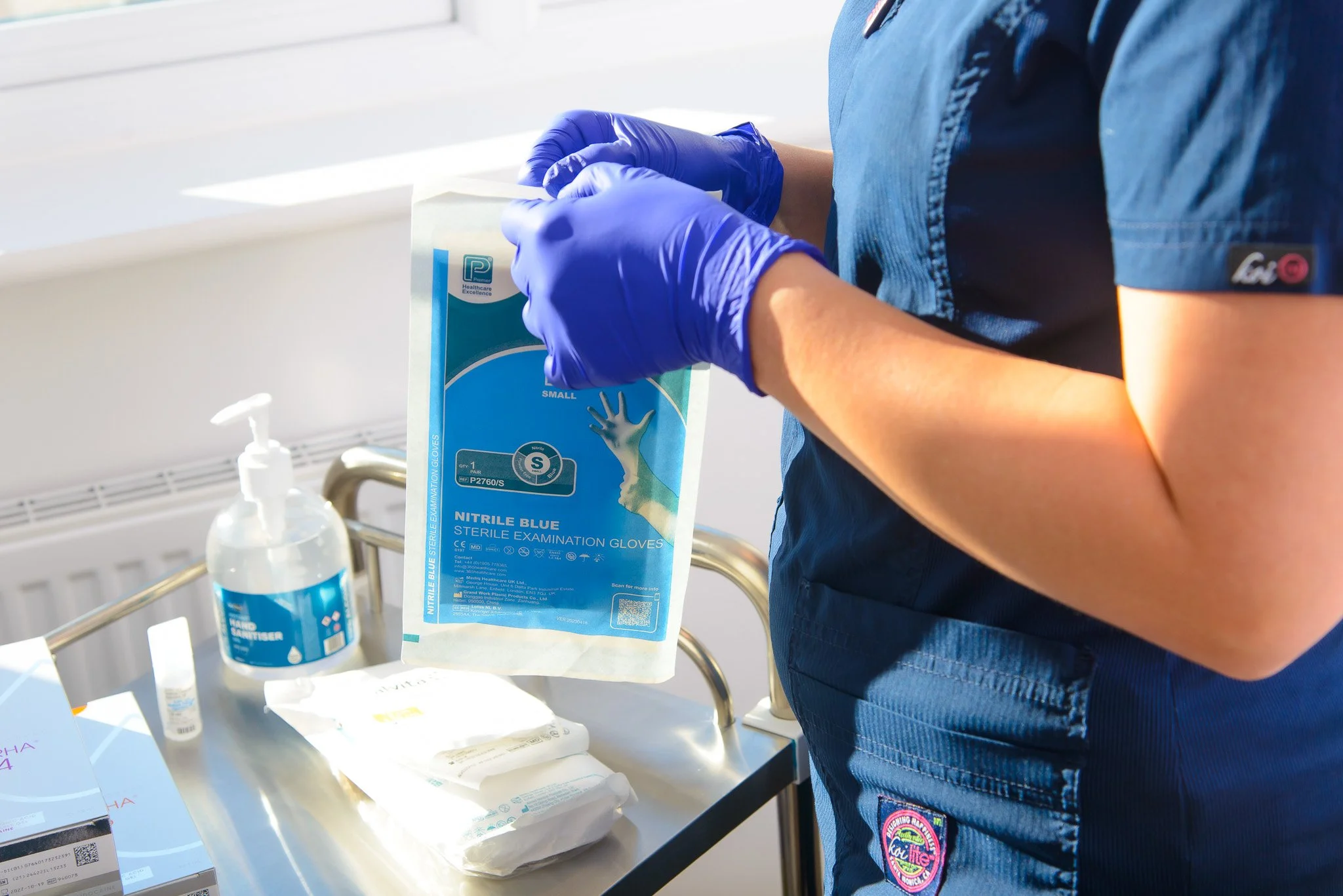 Dr Victoria Waight wearing blue gloves holding a package of nitrile blue sterile examination gloves in a medical setting.