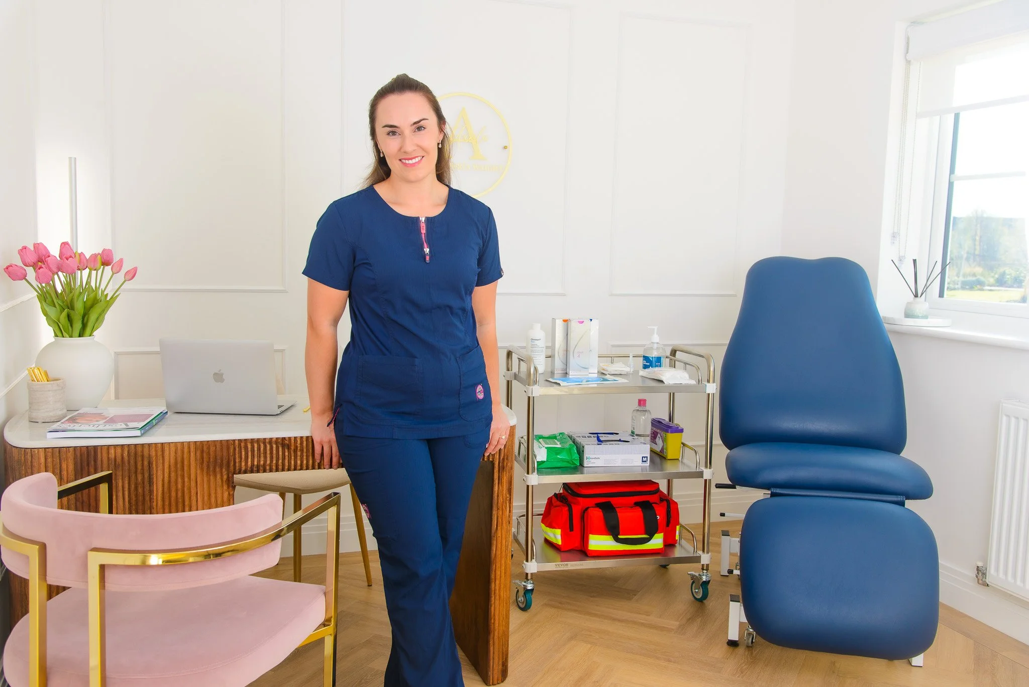 Dr Victoria Waight in blue scrubs standing in aesthetics clinic with a pink chair, a desk with a laptop and flowers, a blue treatment chair, and medical supplies on a cart.
