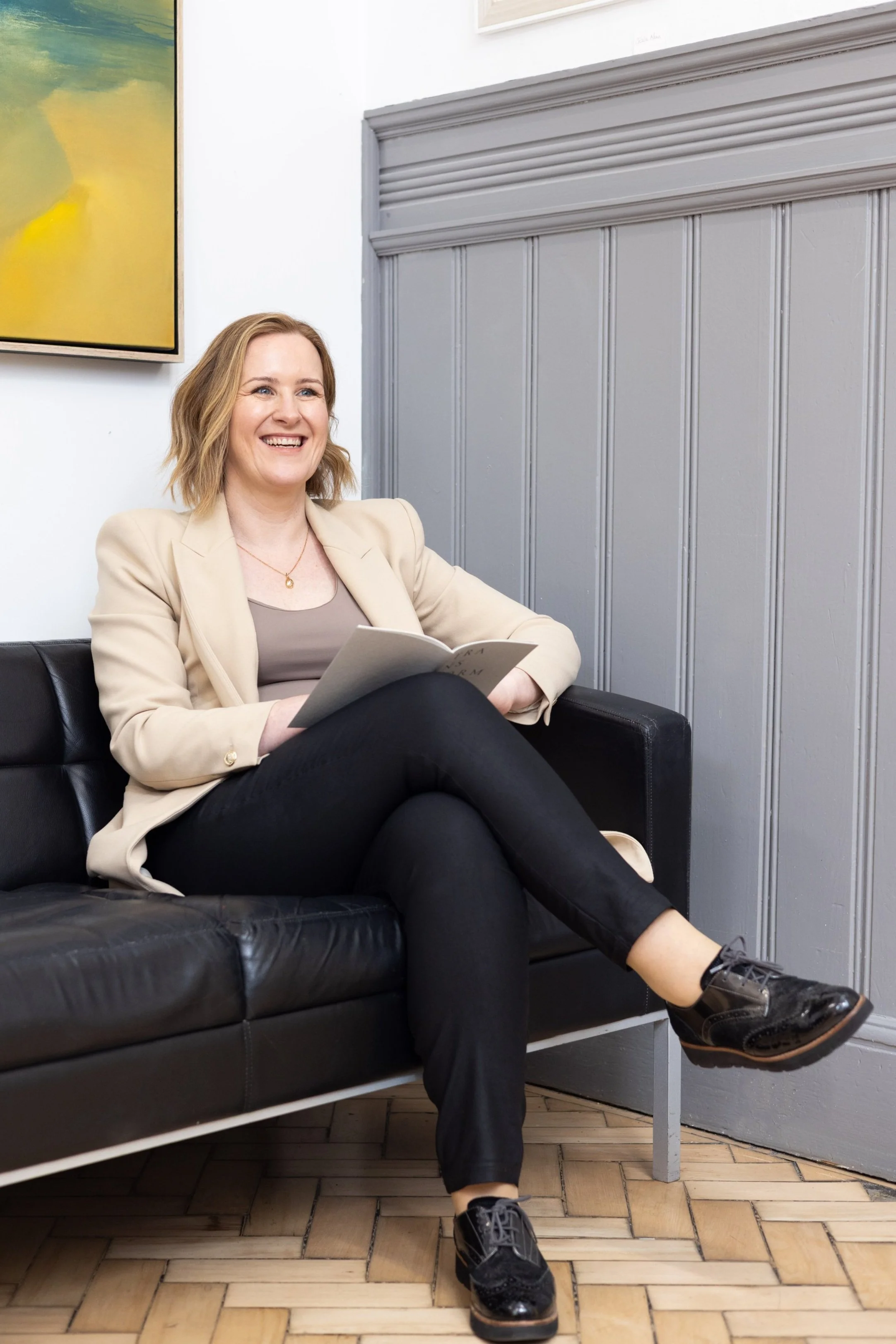 Jill McAlpine, founder of inpurpose, sitting on a black sofa, smiling happily while holding a book or notebook, in a modern indoor space with artwork on the wall.