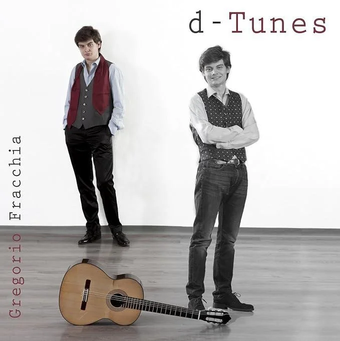 Two young men stand against a plain white wall. The man on the left wears a white shirt with rolled-up sleeves, a vest, and a red scarf. The man on the right wears a white shirt with rolled-up sleeves, a polka-dot vest, and jeans, with arms crossed and smiling. An acoustic guitar lies on the floor in front of them. The wall has text: "d-Tunes" at the top, "Fraccia" vertically on the left, and "Greborto" vertically on the bottom left.