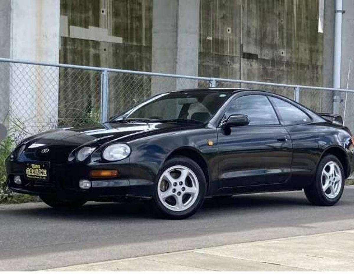 Black Toyota sports car parked on the side of the road near a chain-link fence and concrete wall.
