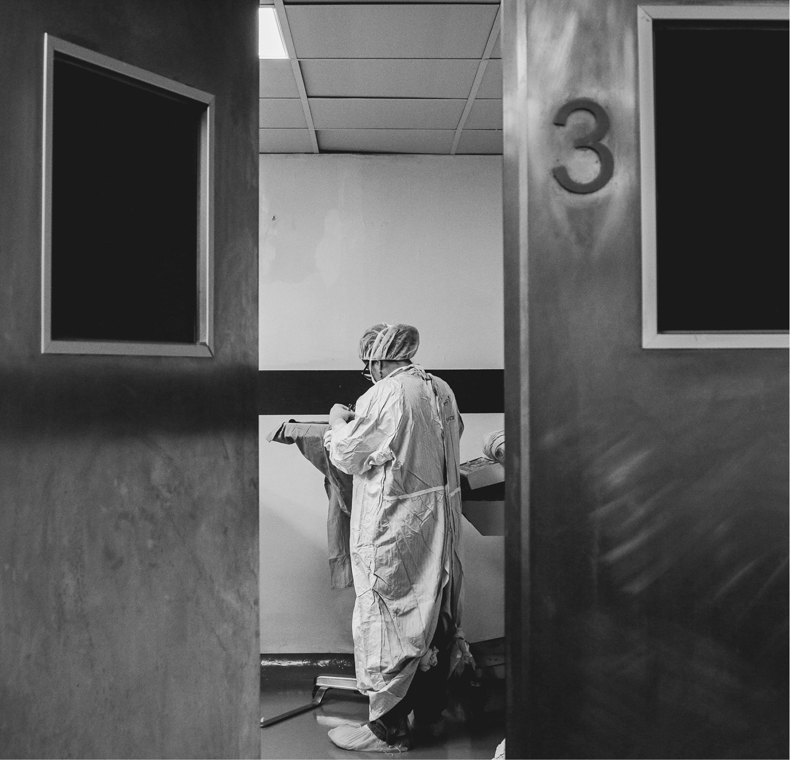 A person dressed in protective medical gear, including a gown, hair cover, mask, and gloves, standing in a clinical setting behind a partially open door marked with the number 3.