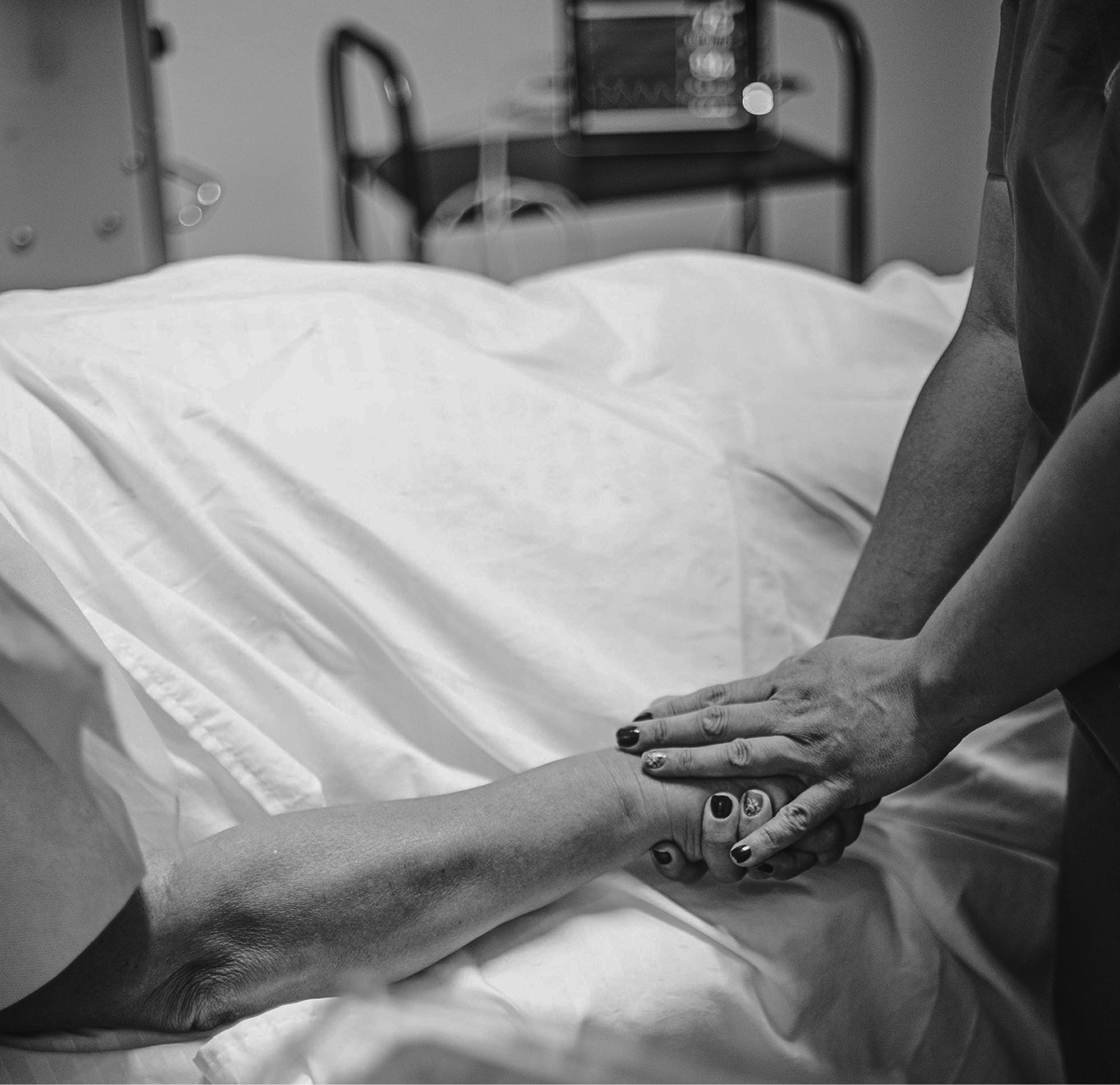 A person in a hospital bed holding hands with another person, possibly a healthcare worker or loved one, in a medical setting.