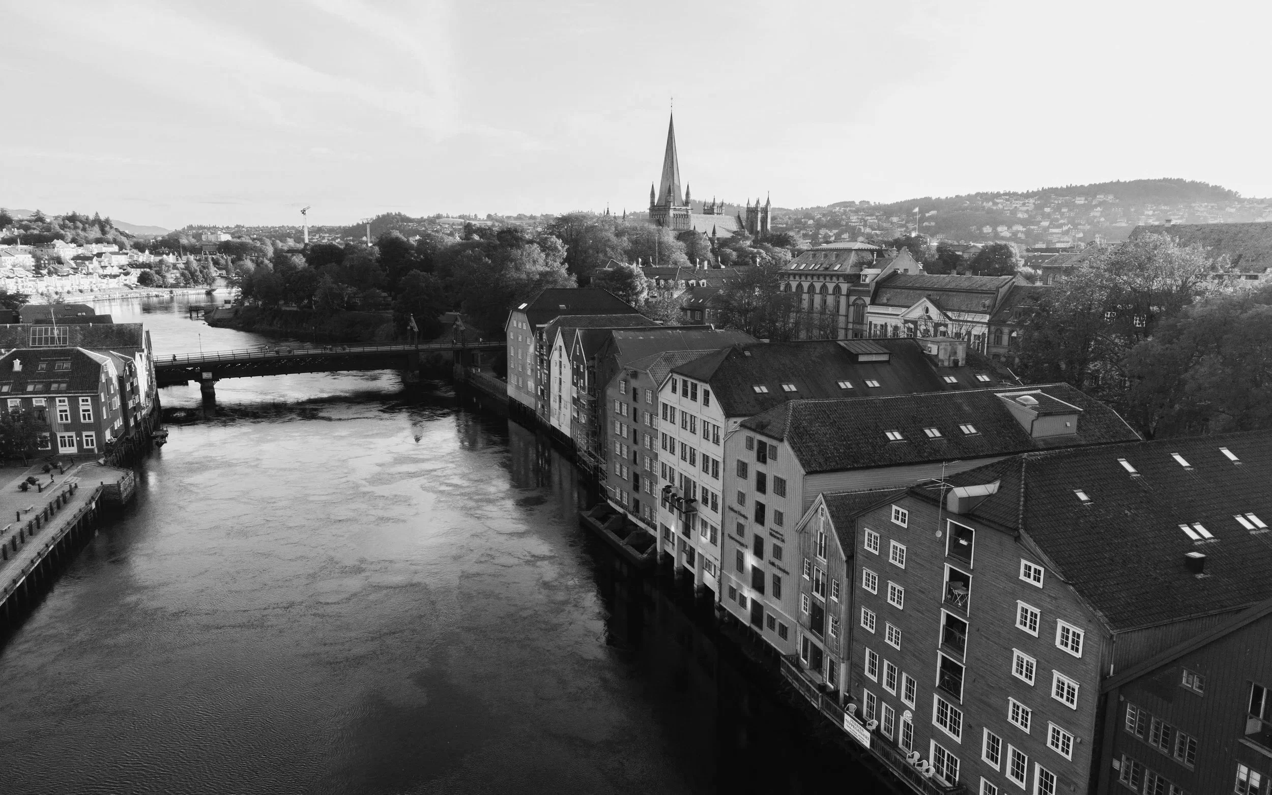 Black and white photograph of a scenic cityscape featuring a river with boats, a bridge, historic buildings along the waterfront, and a prominent cathedral with a tall spire in the background.