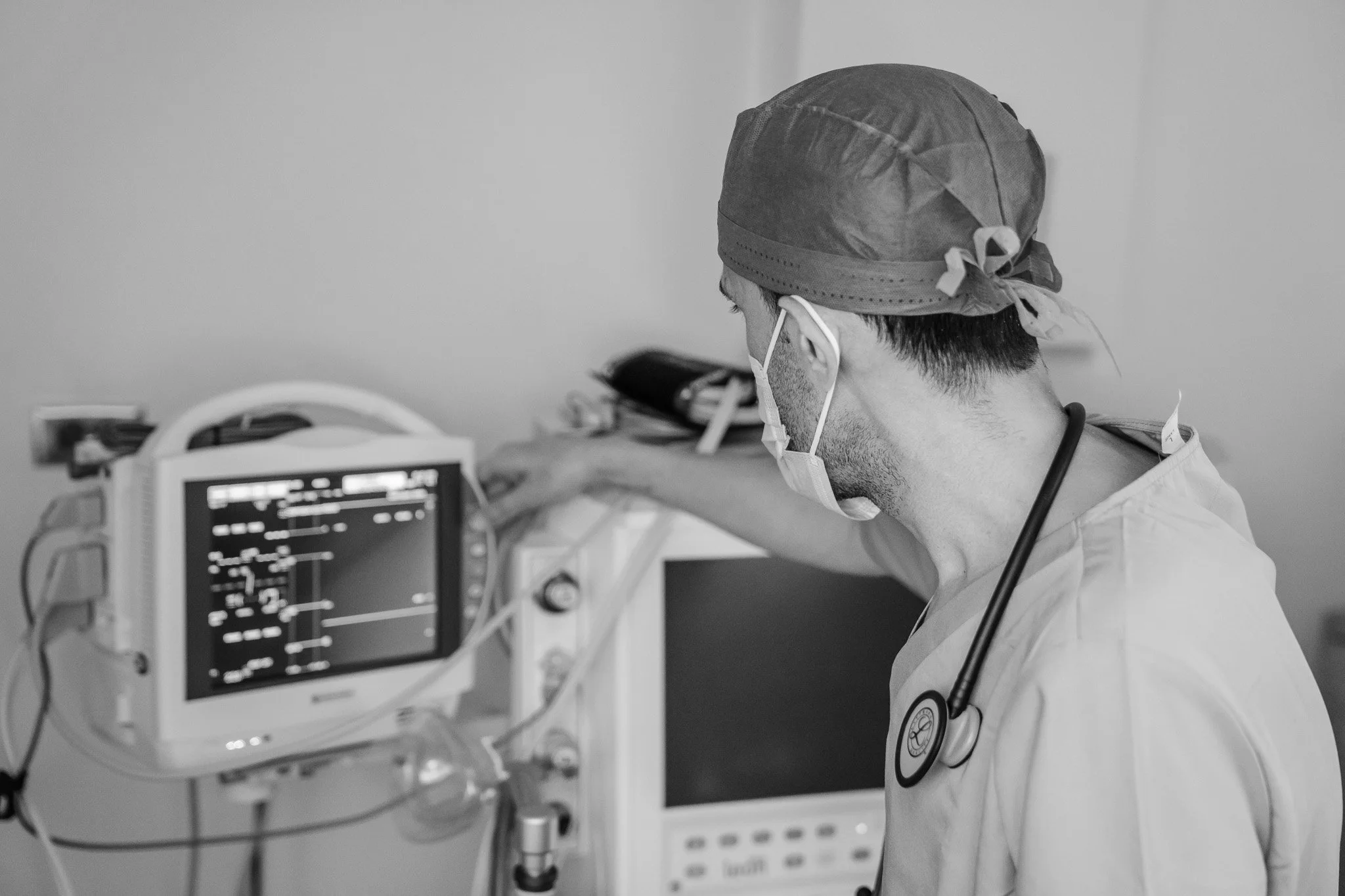 A medical professional wearing a surgical cap and face mask working in a hospital, looking at a medical monitor displaying vital signs.